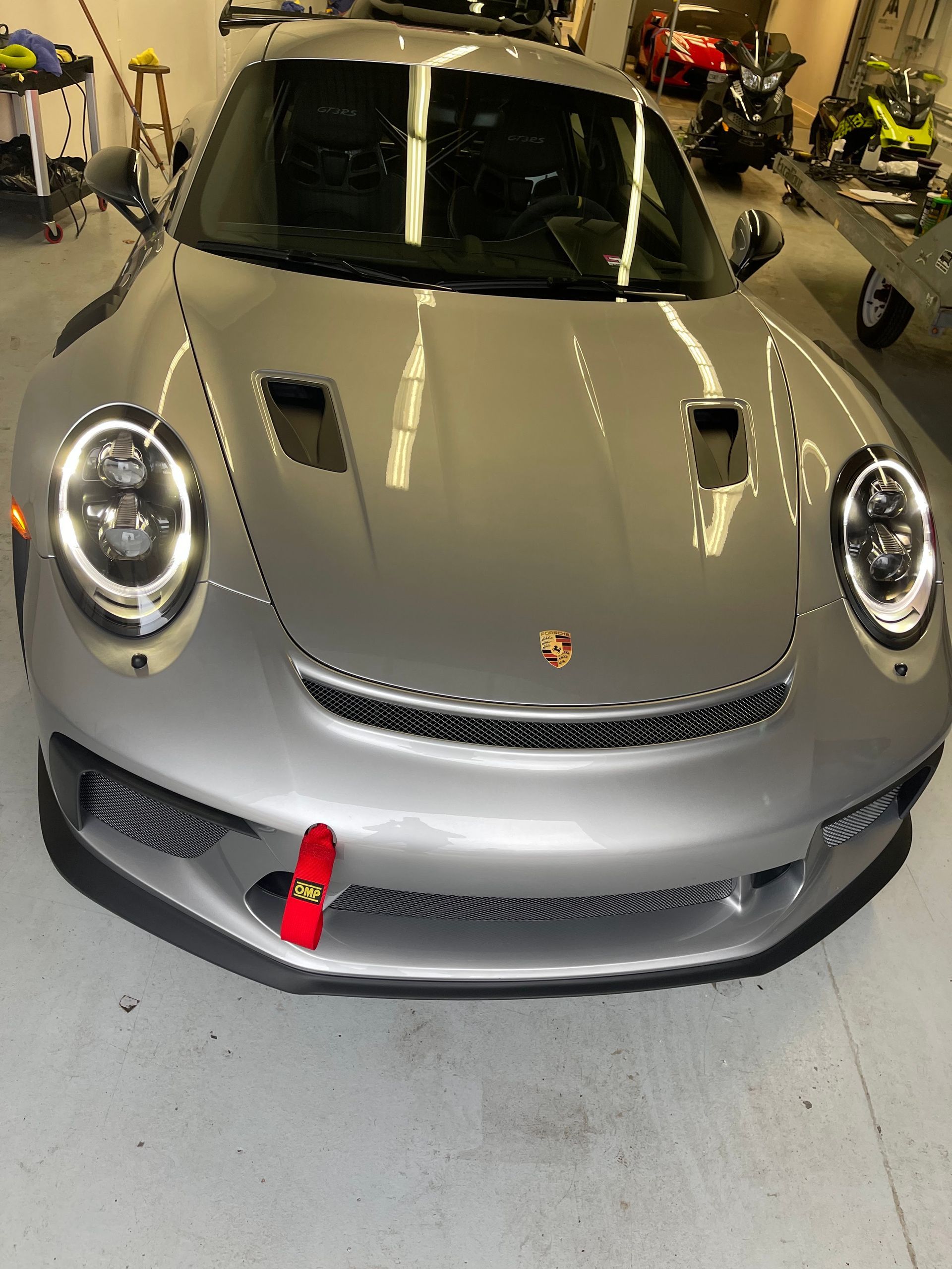 Silver Porsche 911 GT3 in a garage, featuring a front red tow strap, LED headlights, and hood vents.