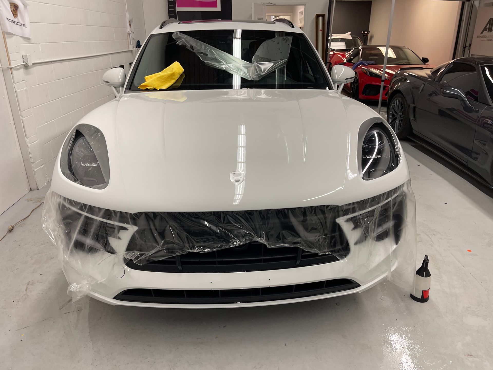 A white Porsche with protective plastic film over the windshield and grille in a garage with other cars.