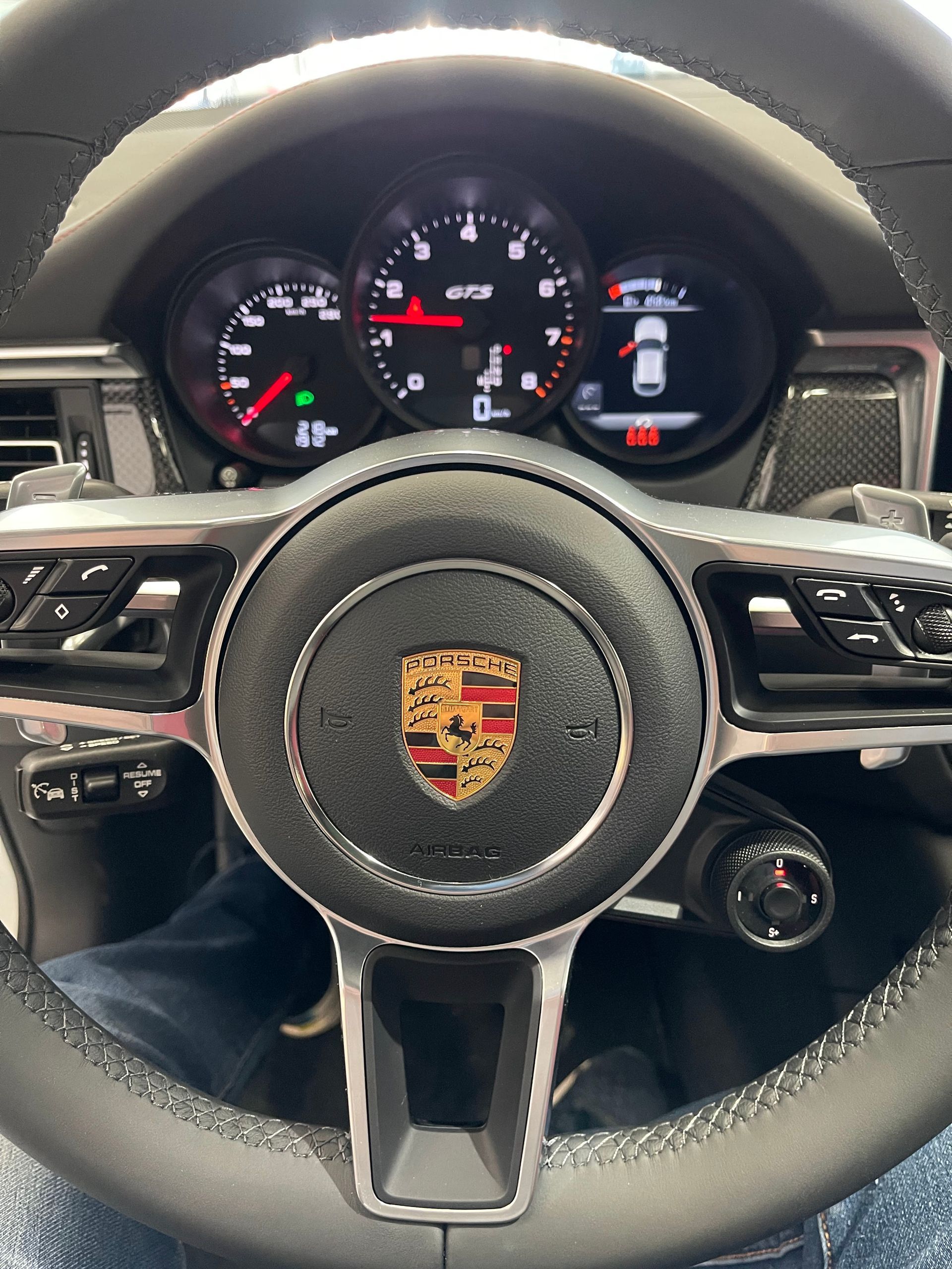 Driver's view of a Porsche steering wheel with a central emblem and instrument cluster showing dials and digital data.