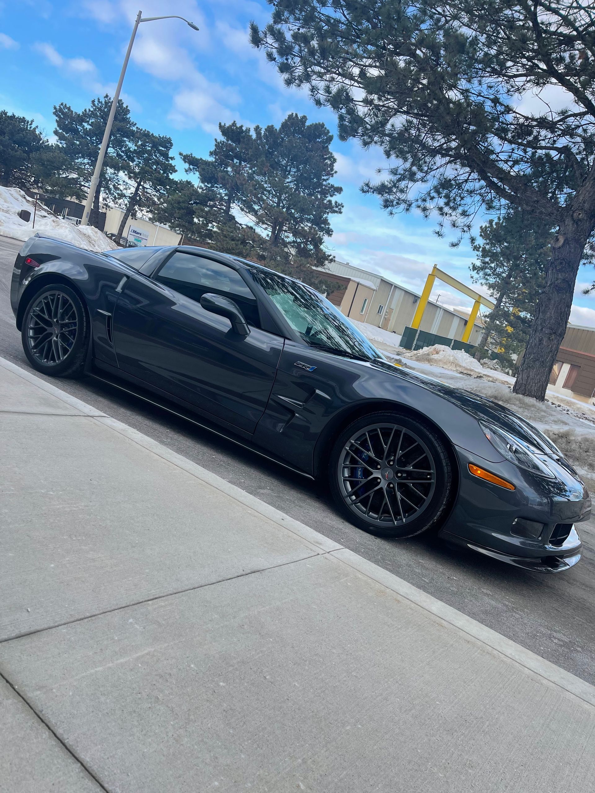 A dark gray Chevrolet Corvette parked on a snowy roadside with bare trees in the background under a blue sky.