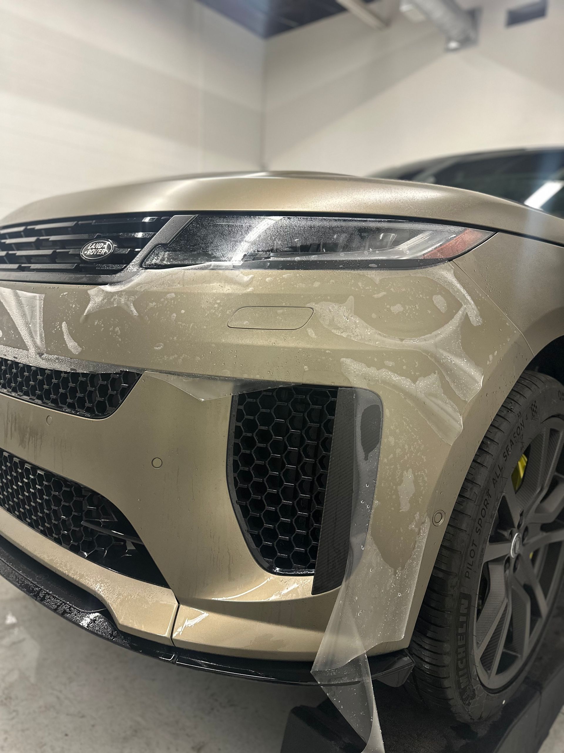A gold Range Rover in a shop with a protective plastic film being applied to the front bumper.