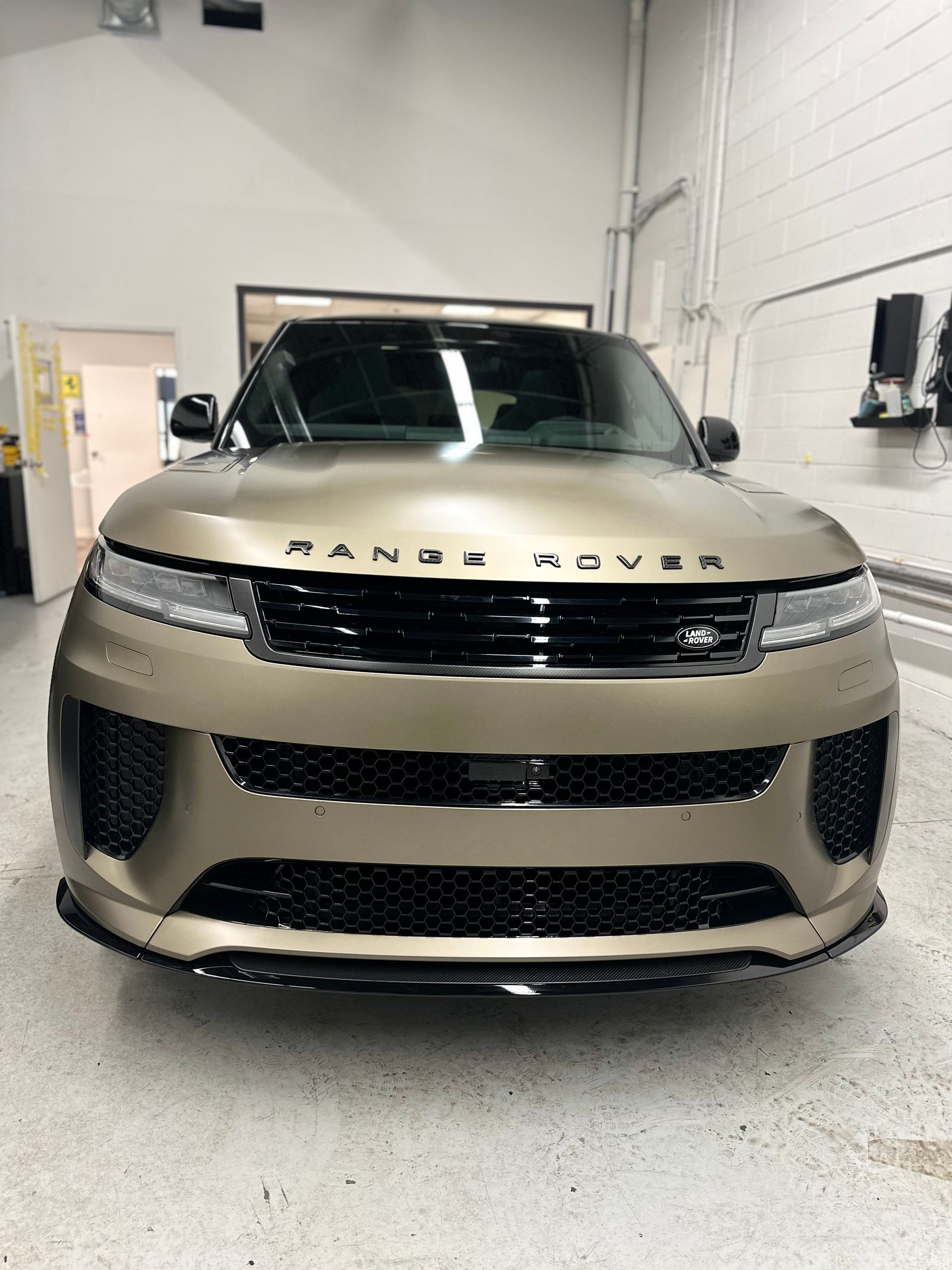 A front-facing view of a gold-colored Range Rover parked inside a brightly lit automotive workshop.