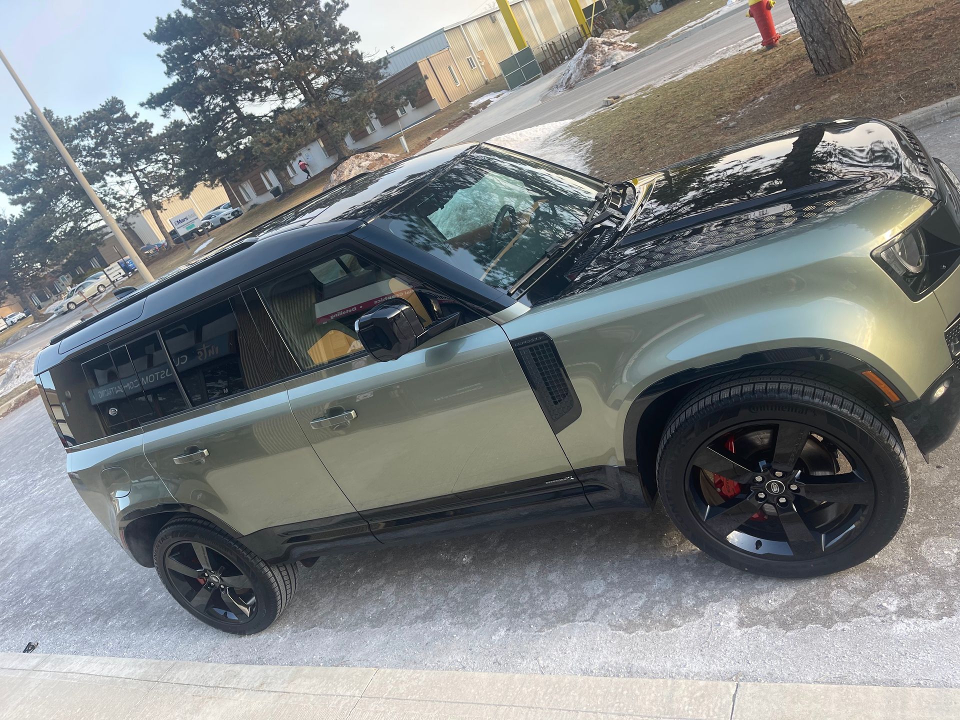 An olive-green Land Rover Defender parked on an asphalt surface, featuring a black roof, black trim, and black wheels.