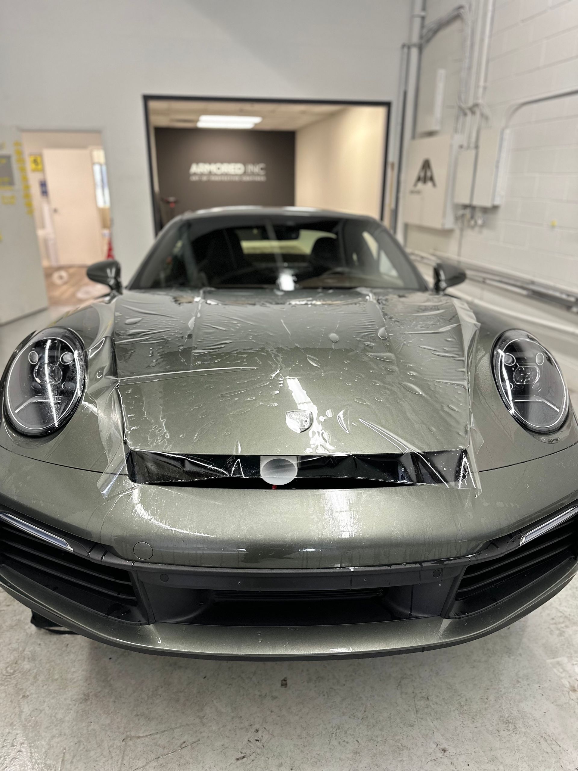 A grey Porsche 911 being applied with protective paint film in an automotive garage.