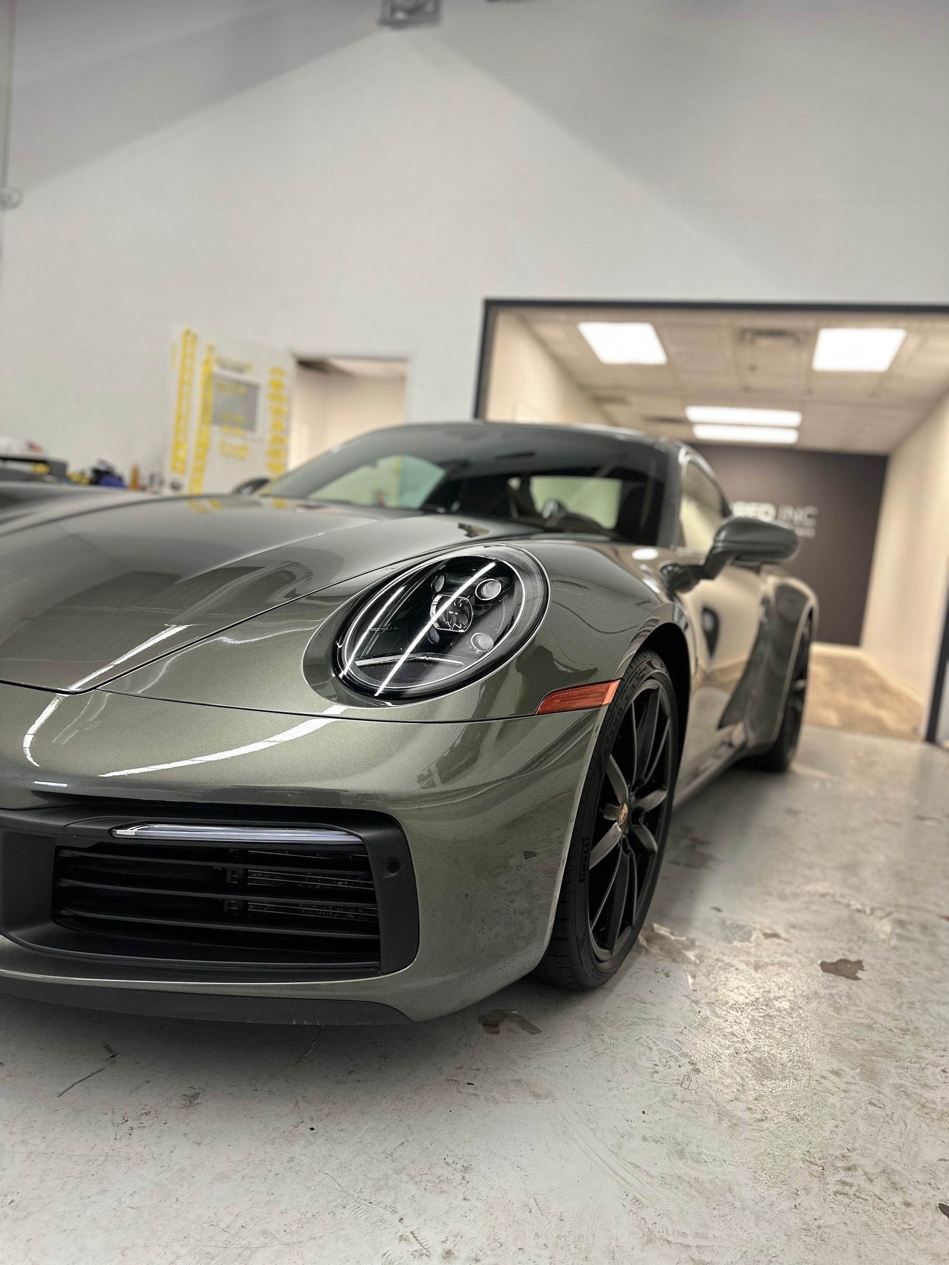 A sleek metallic olive-green Porsche 911 parked inside a brightly lit professional garage.