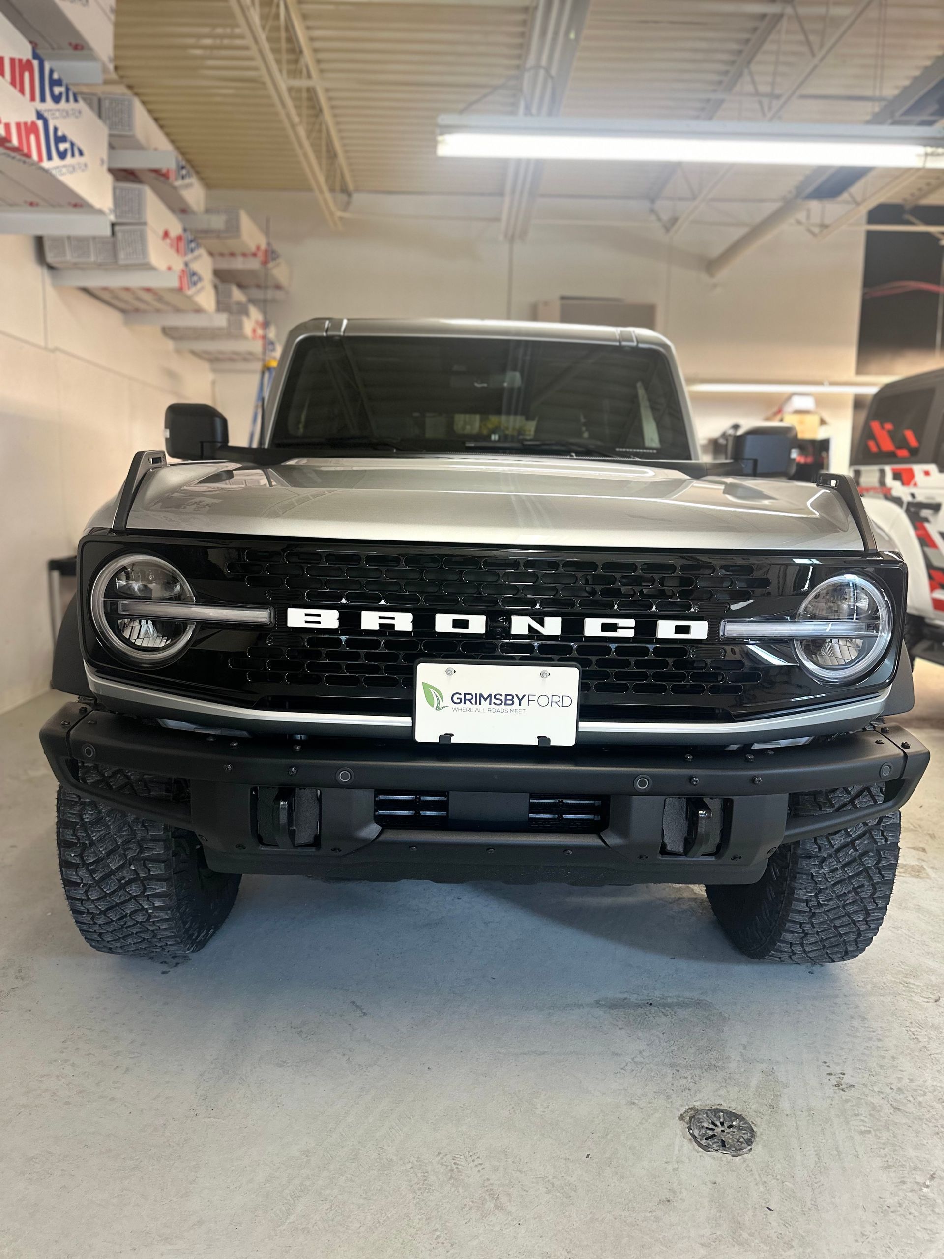 A silver Ford Bronco parked inside a garage, featuring a black grille, large headlights, and a sturdy front bumper.