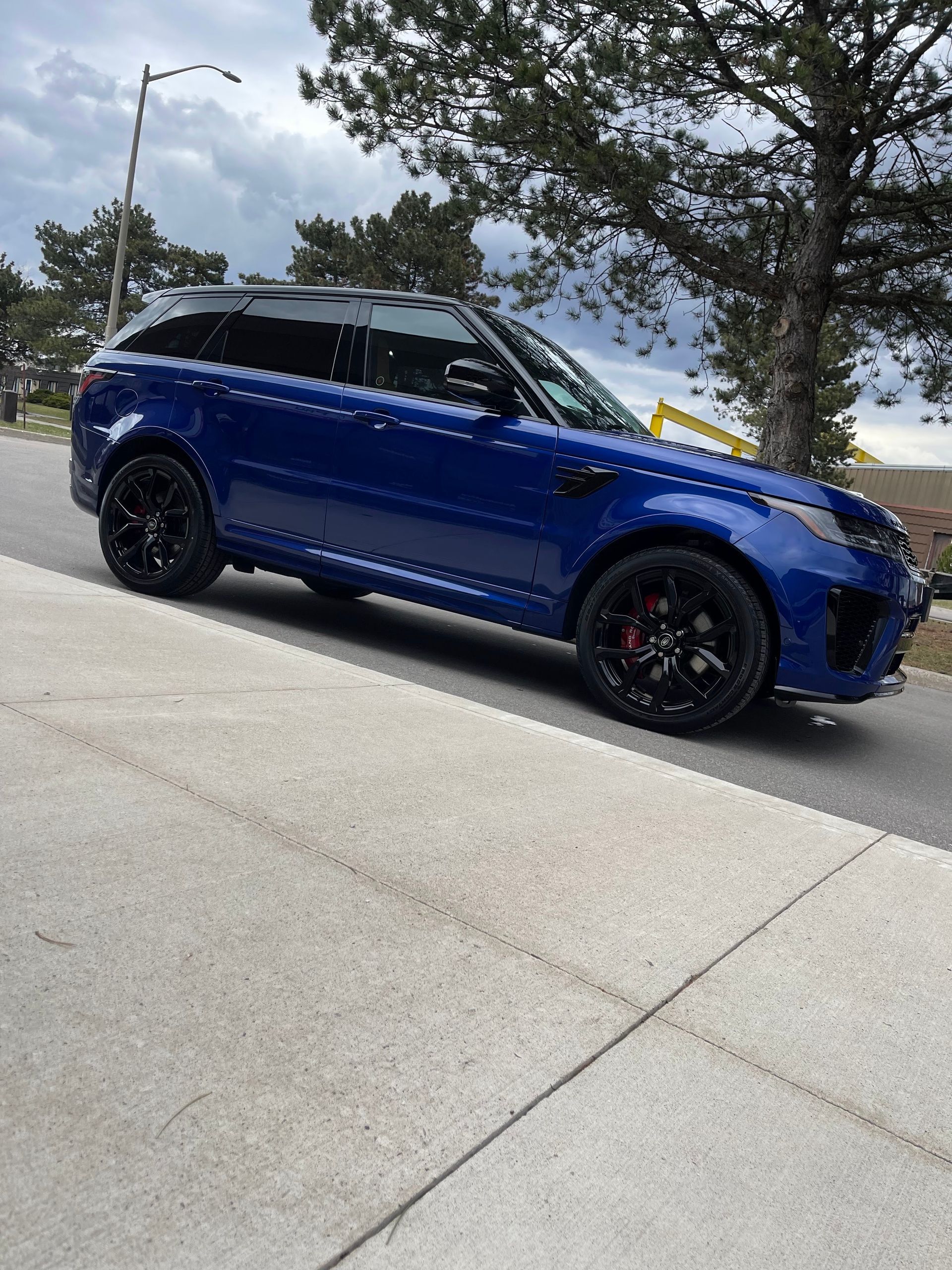 A blue Land Rover Range Rover parked on a paved street next to a concrete sidewalk, with trees and a cloudy sky above.