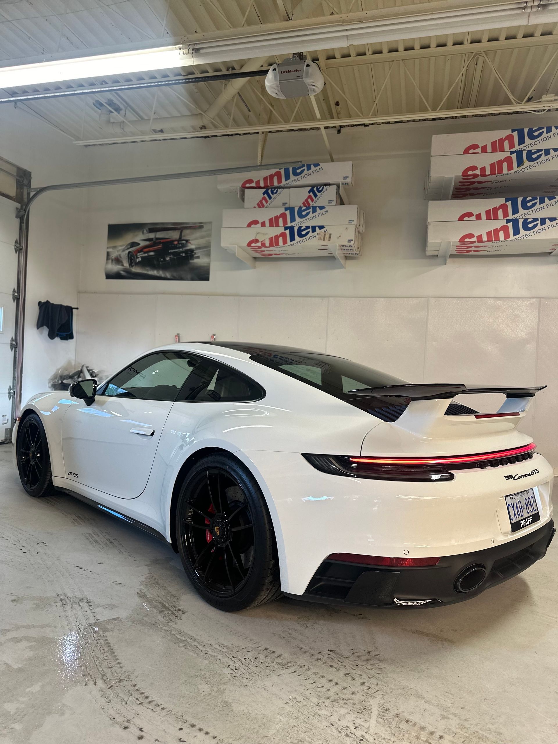 A white Porsche 911 GT3 with a rear wing and black wheels, parked inside a garage with film supply boxes on the wall.