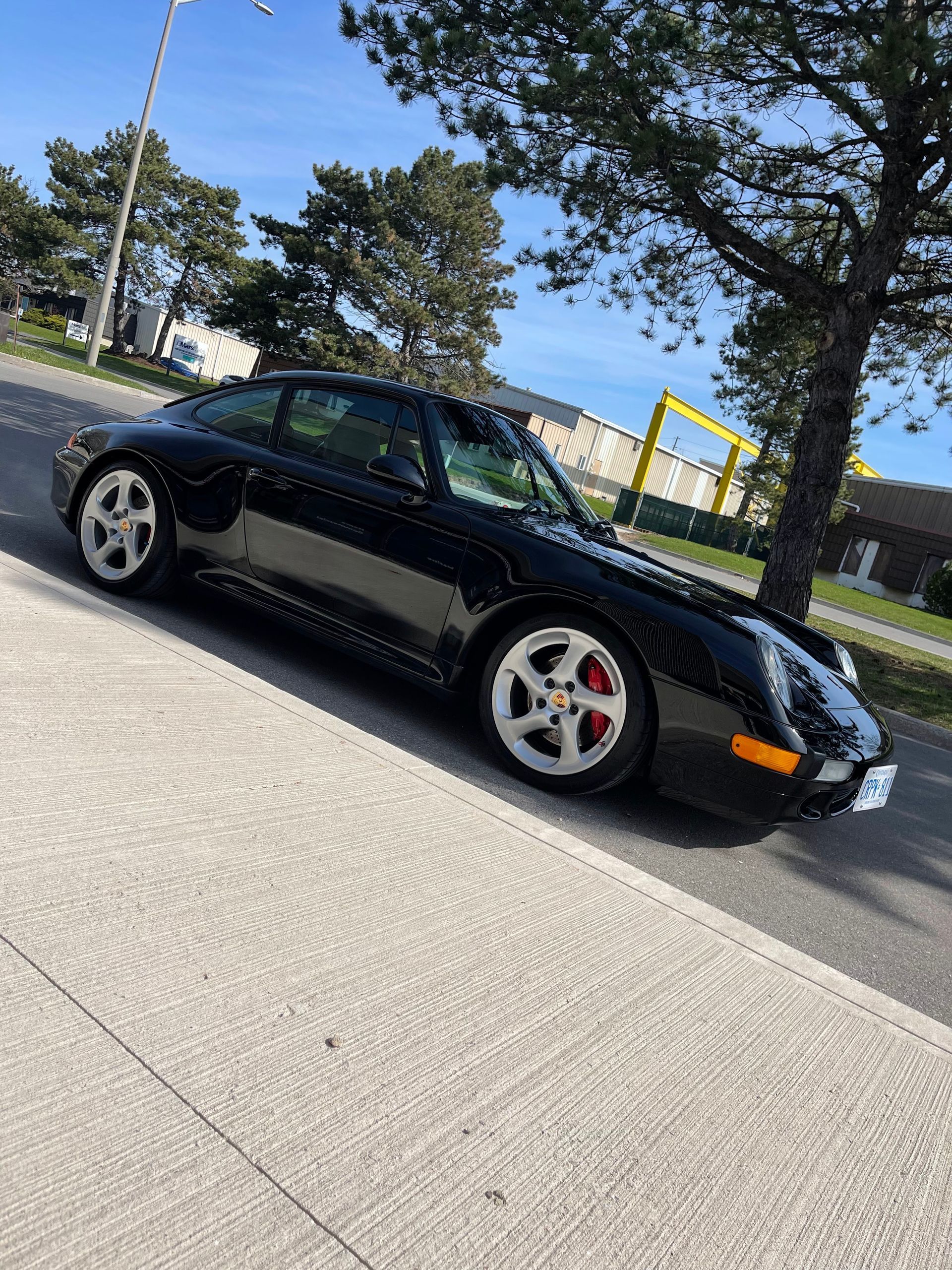 A black Porsche 911 parked on the side of a road near a tree on a sunny day.