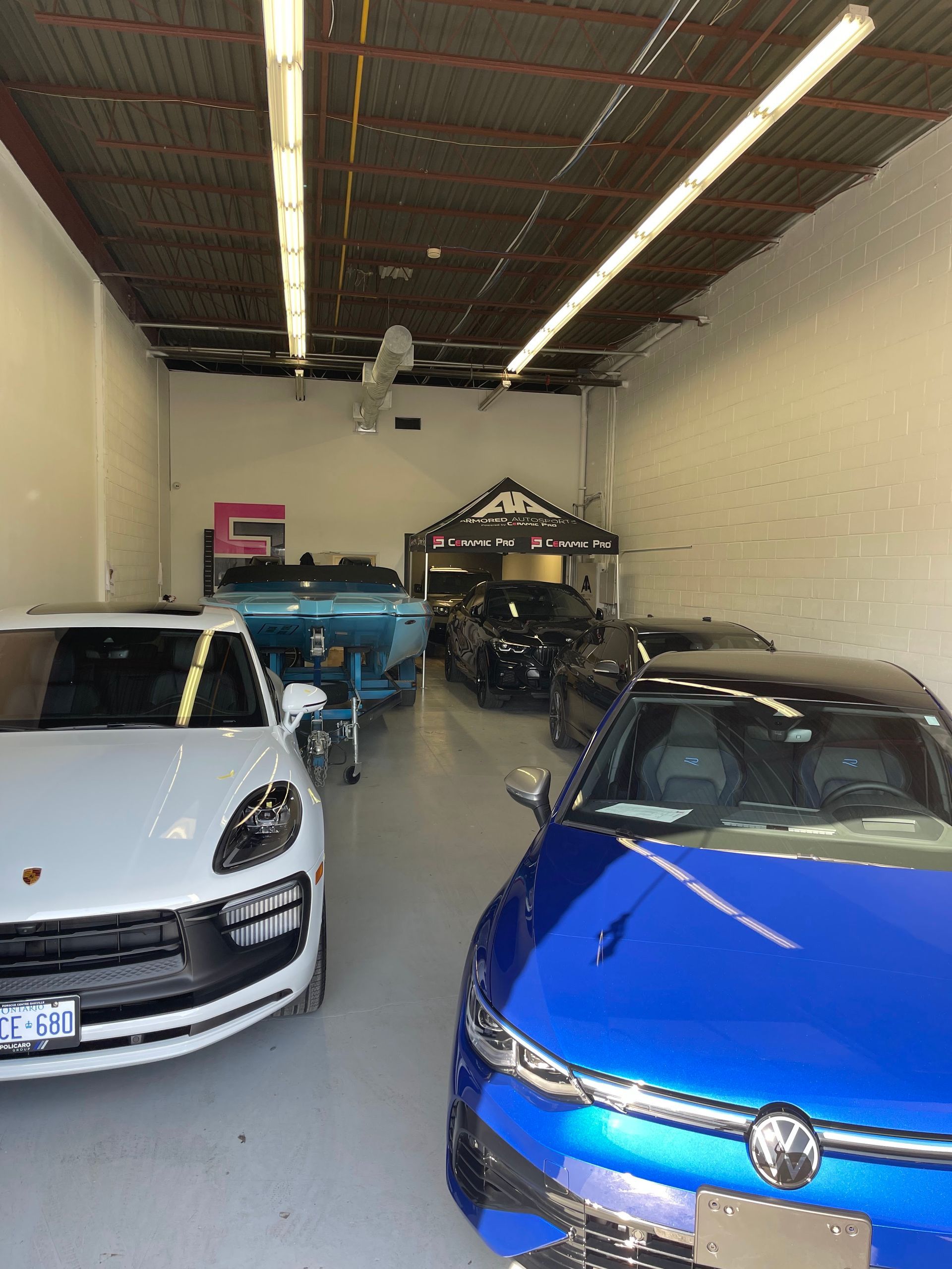 A white Porsche and a blue Volkswagen parked inside a brightly lit automotive workshop with a boat in the background.