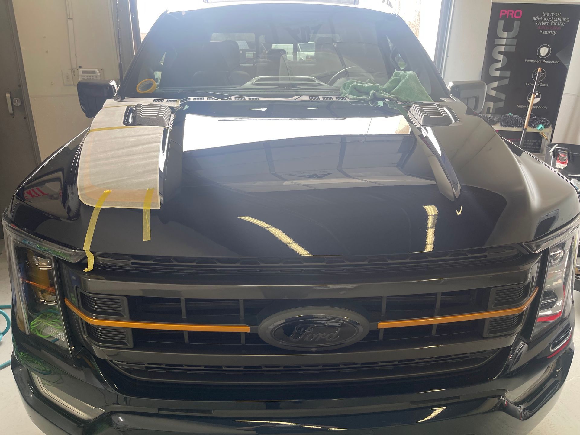Front view of a black Ford pickup truck in a shop, with a protective pad taped to the hood during maintenance work.