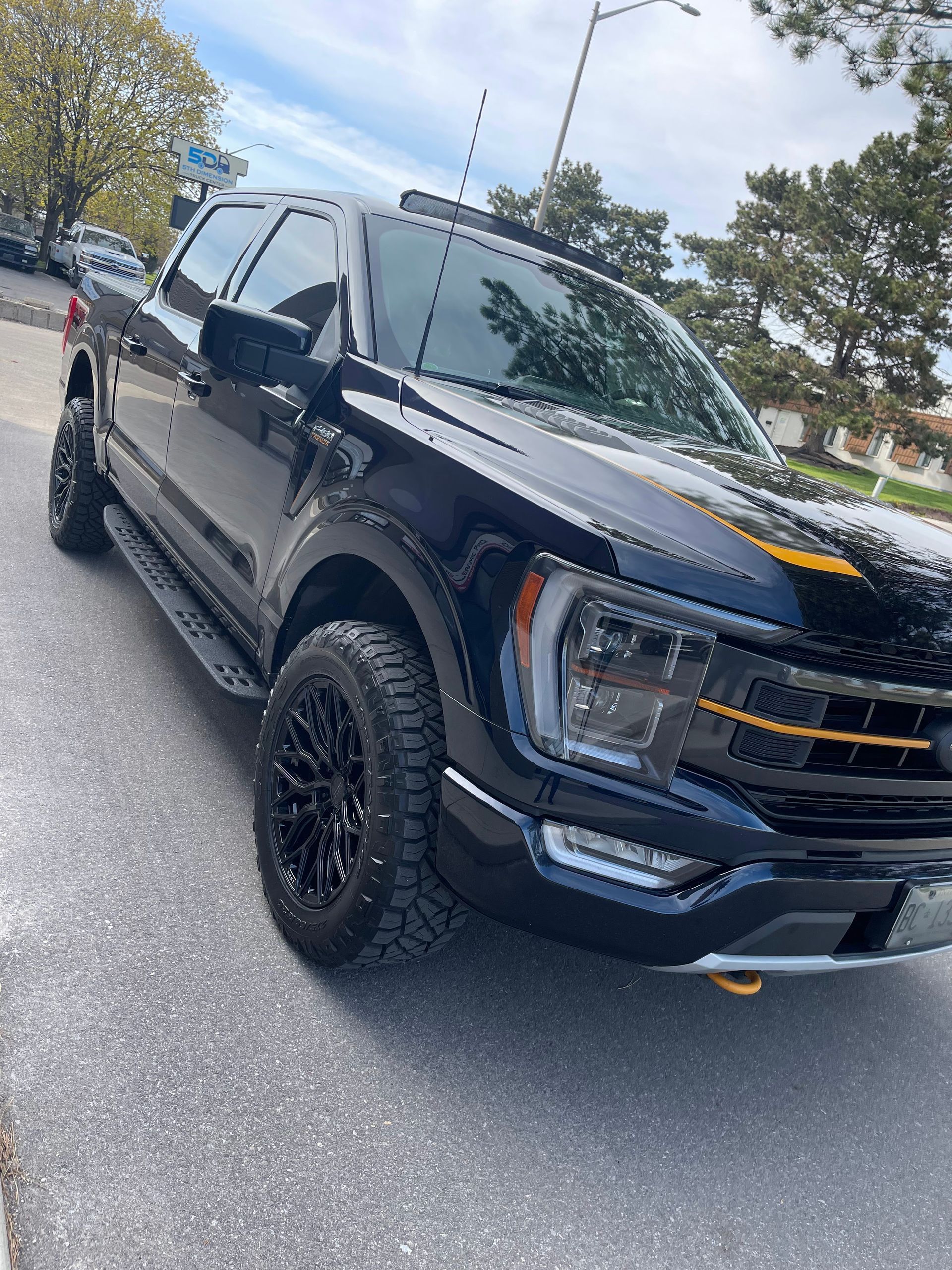 A black Ford F-150 pickup truck with black off-road wheels, amber grille accents, and a light bar, parked on pavement.