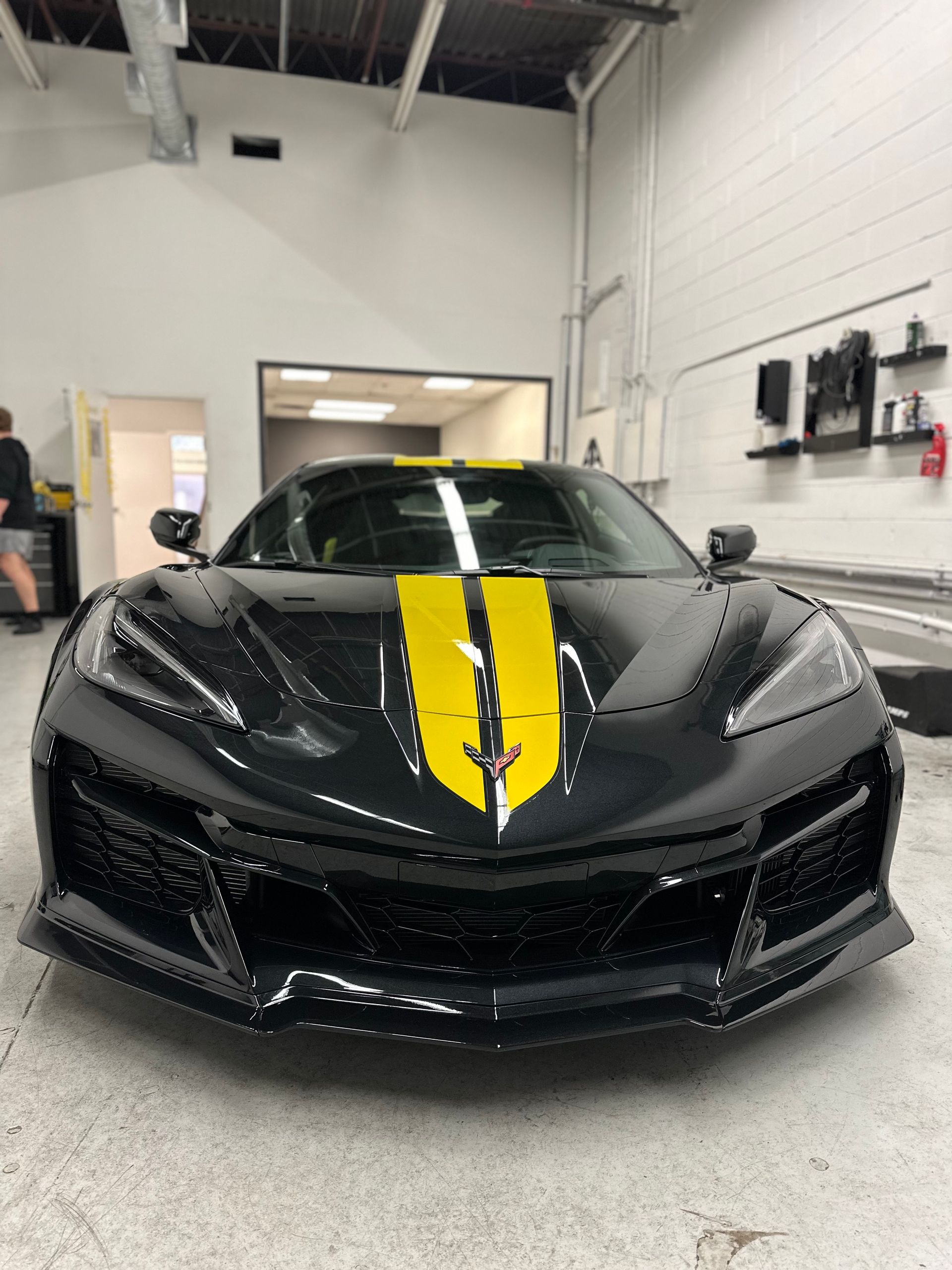 A black Chevrolet Corvette Z06 with a yellow racing stripe parked in a brightly lit automotive workshop.