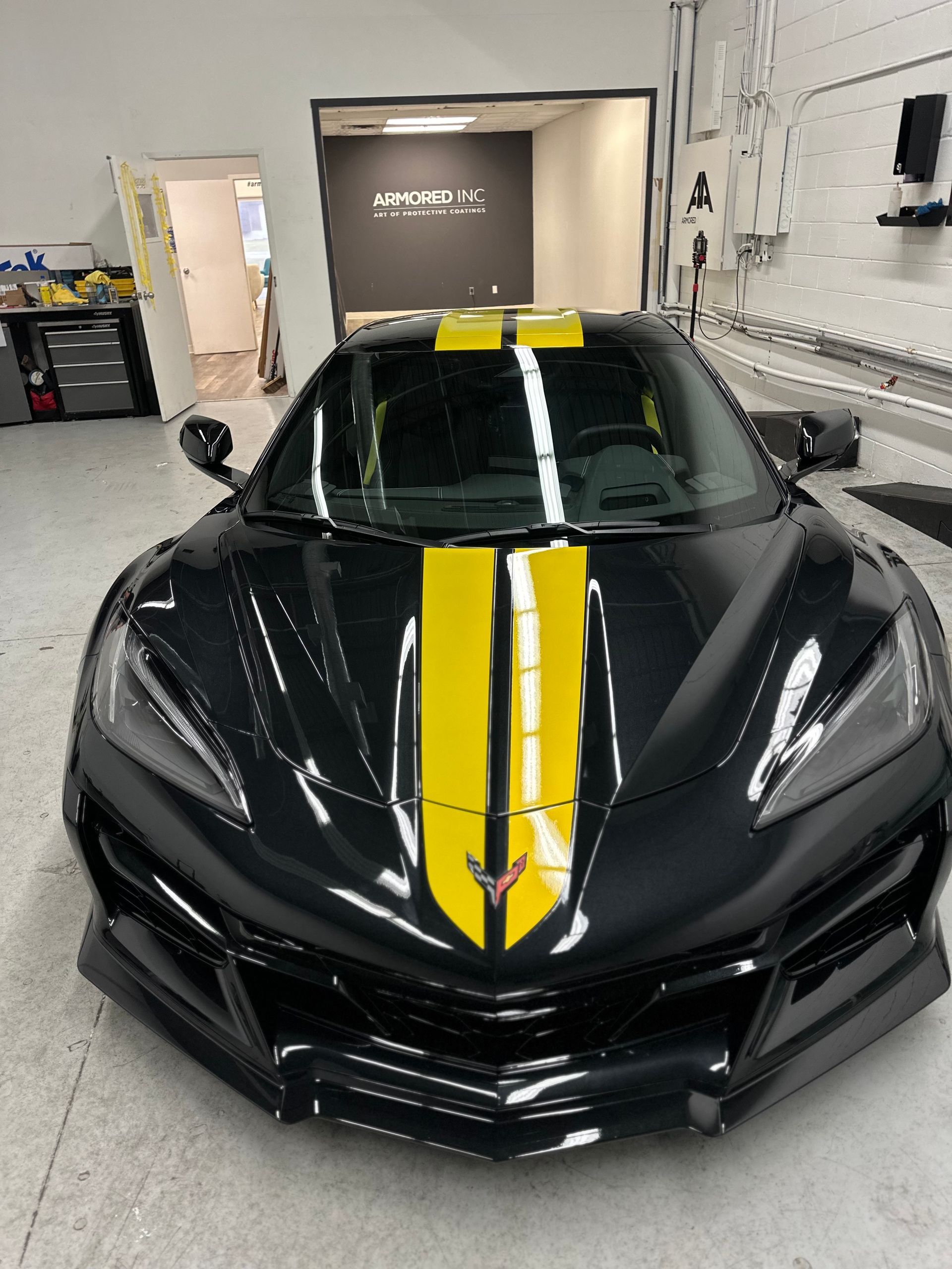 A black Chevrolet Corvette with yellow racing stripes parked inside a bright, clean garage.