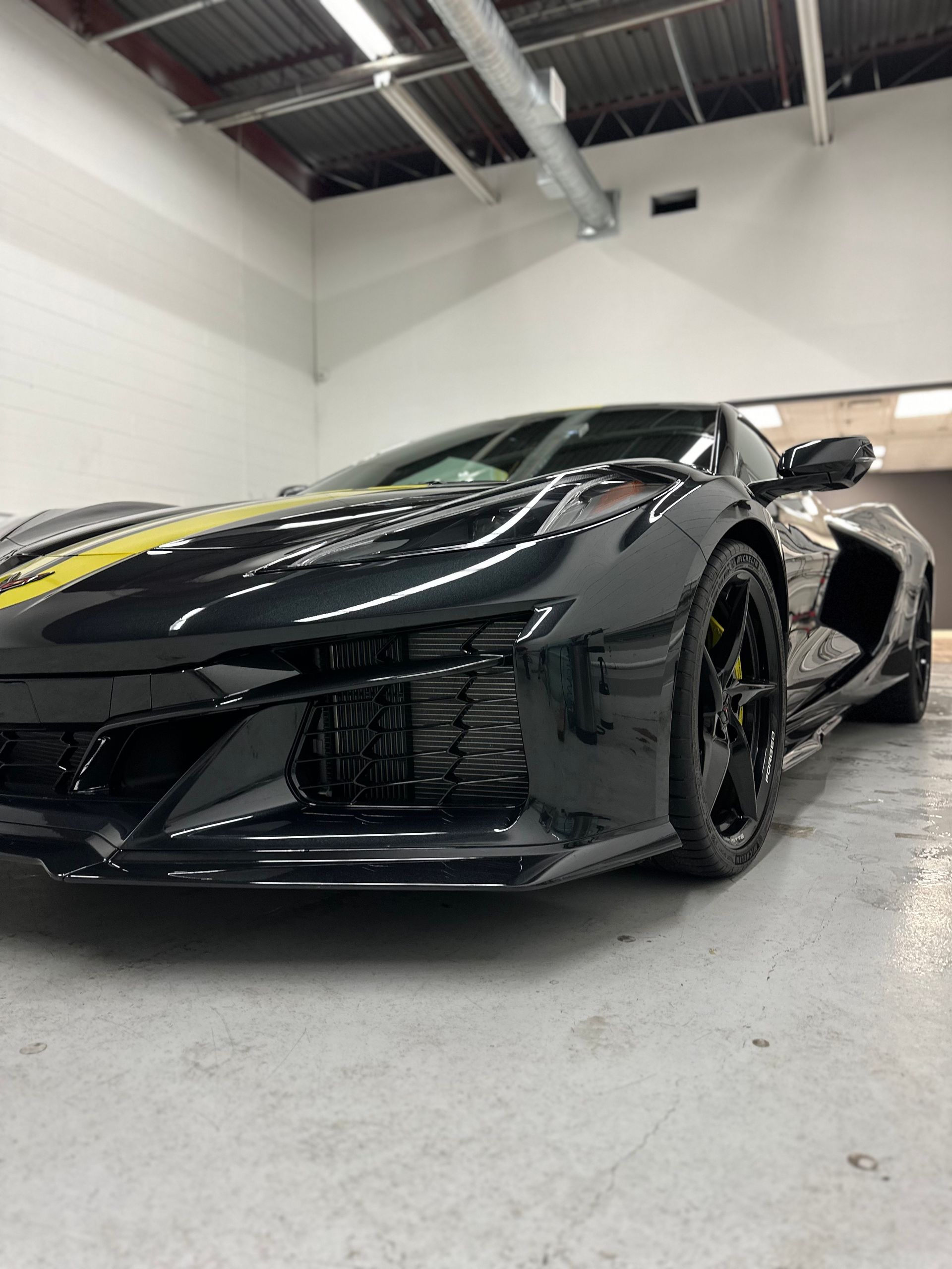 A low-angle shot of a black Chevrolet Corvette with a yellow racing stripe, parked in a well-lit, industrial garage.