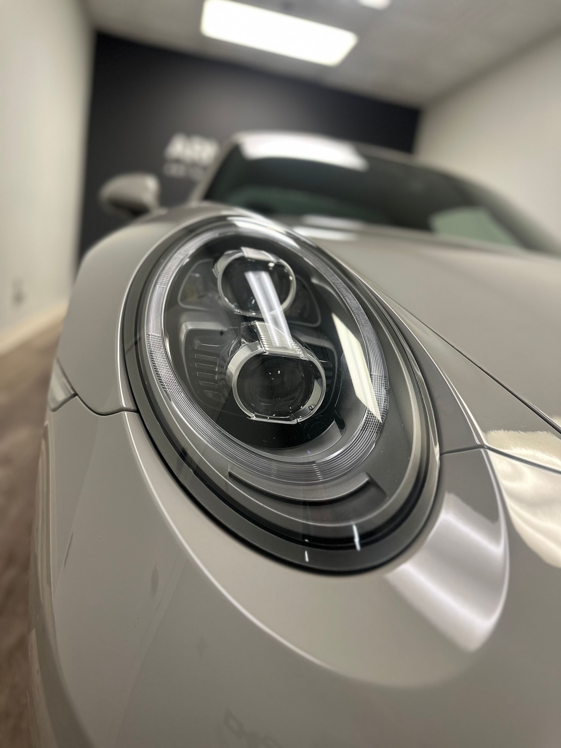 A close-up, angled view of a light gray Porsche 911 headlight in a bright, modern indoor garage.