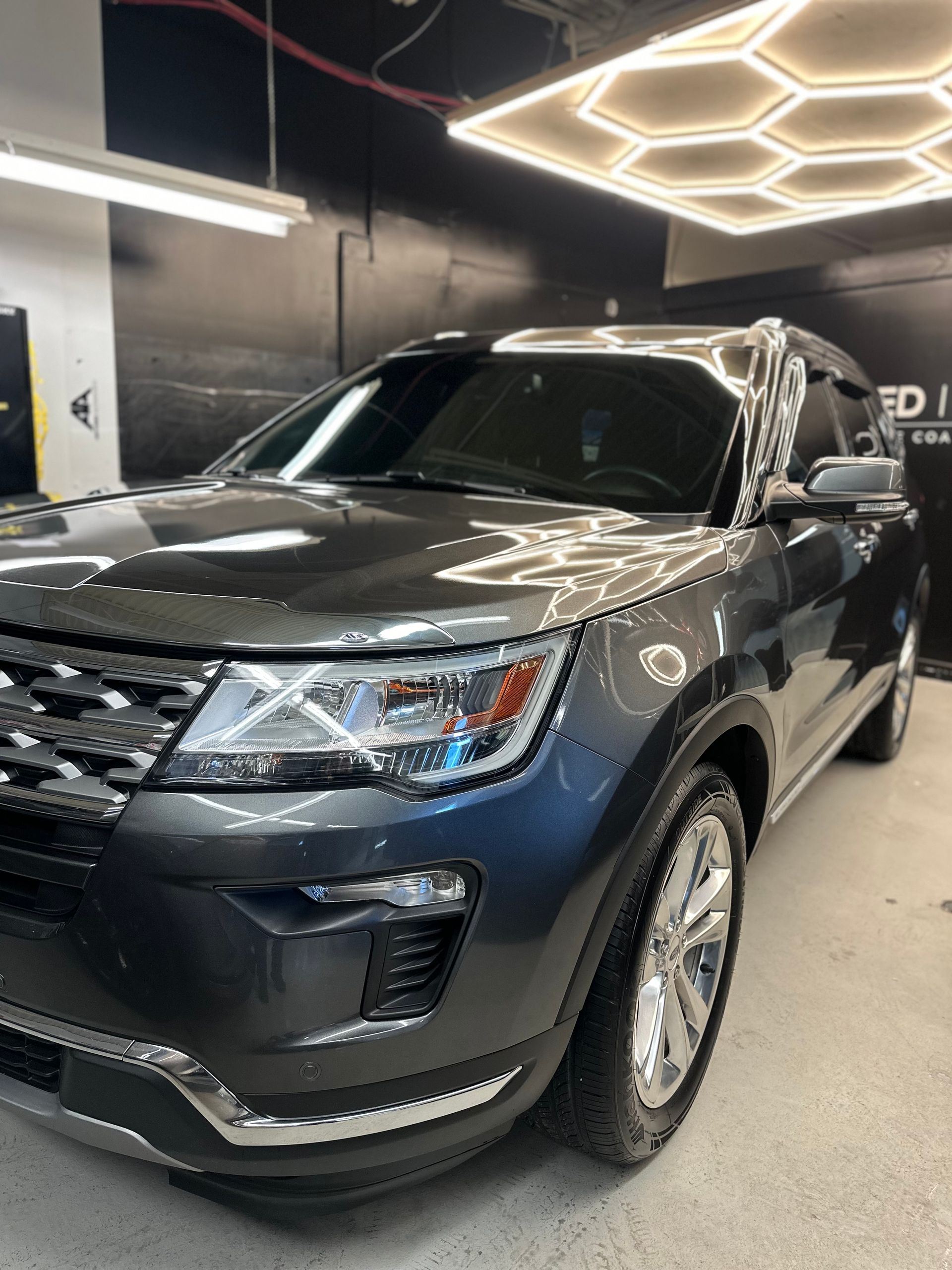 A dark grey Ford Explorer SUV parked inside a workshop with a geometric overhead light panel.