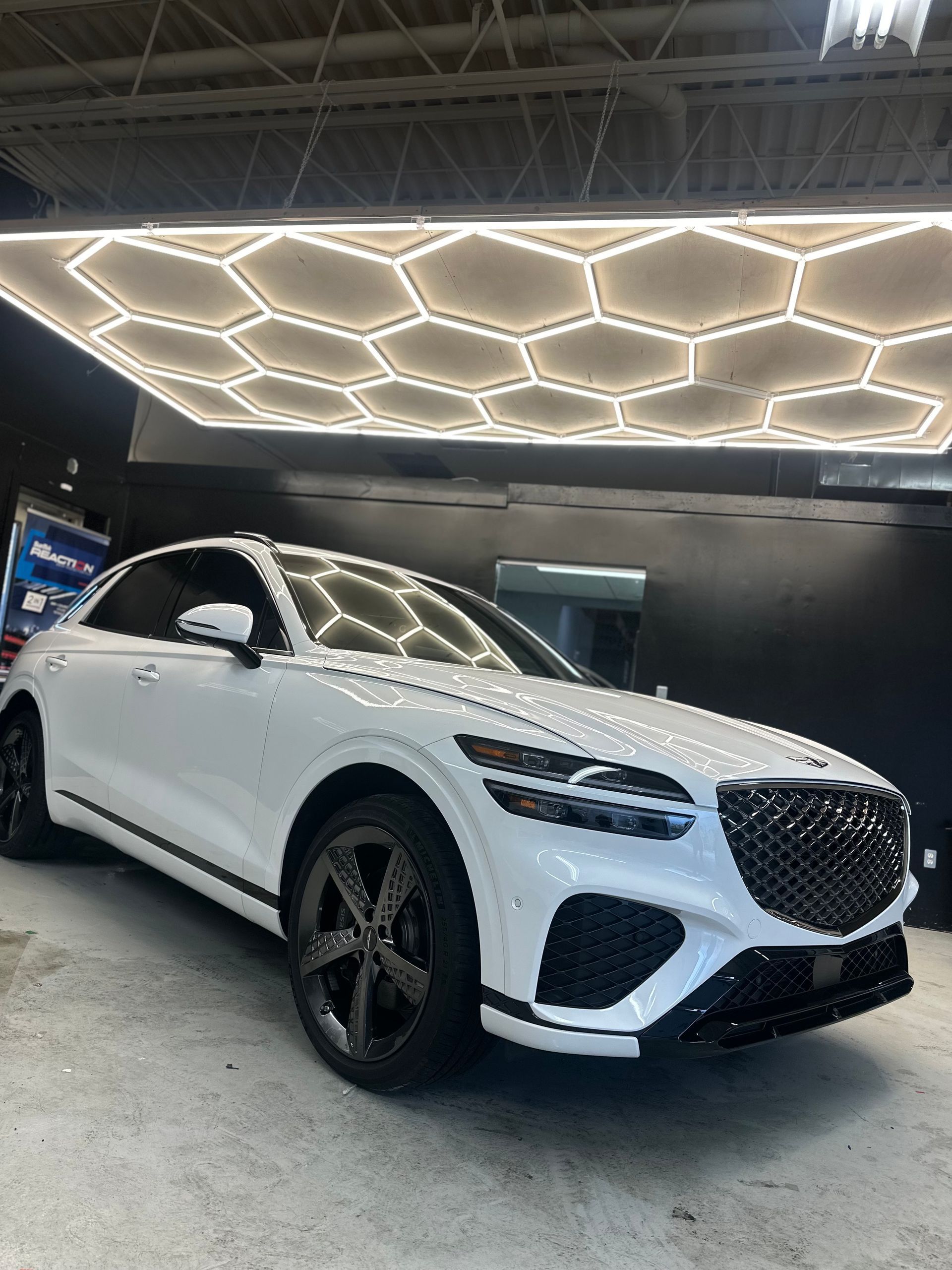 A white Genesis GV70 SUV parked in a garage with a hexagonal LED ceiling light pattern reflecting on its hood.