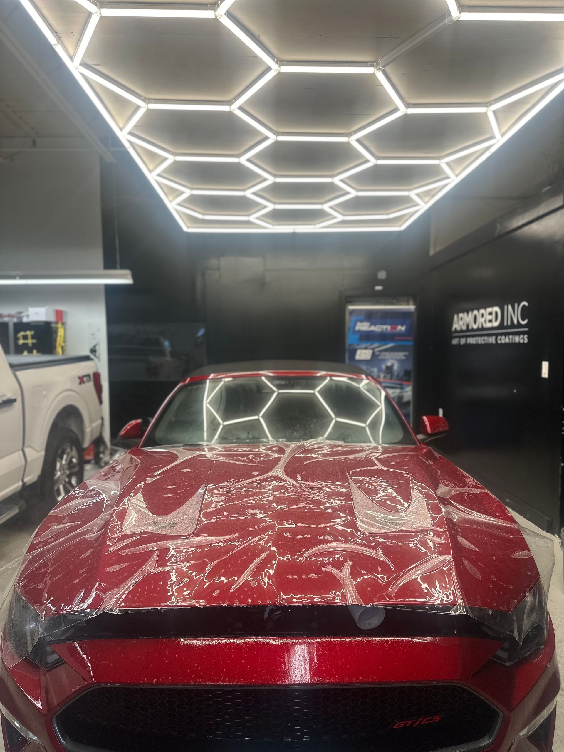 A red Mustang in a garage with honeycomb LED ceiling lights, covered in protective film during a detailing process.