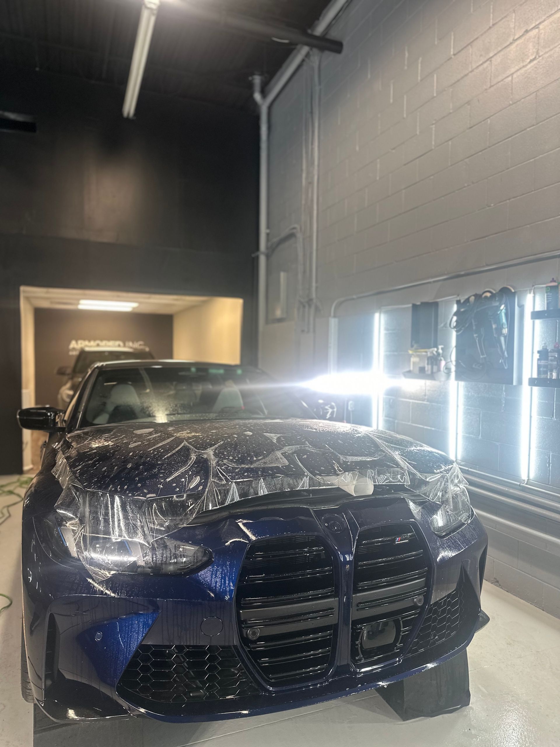 A dark blue car in a bright workshop, partially covered in clear protective film during an installation process.
