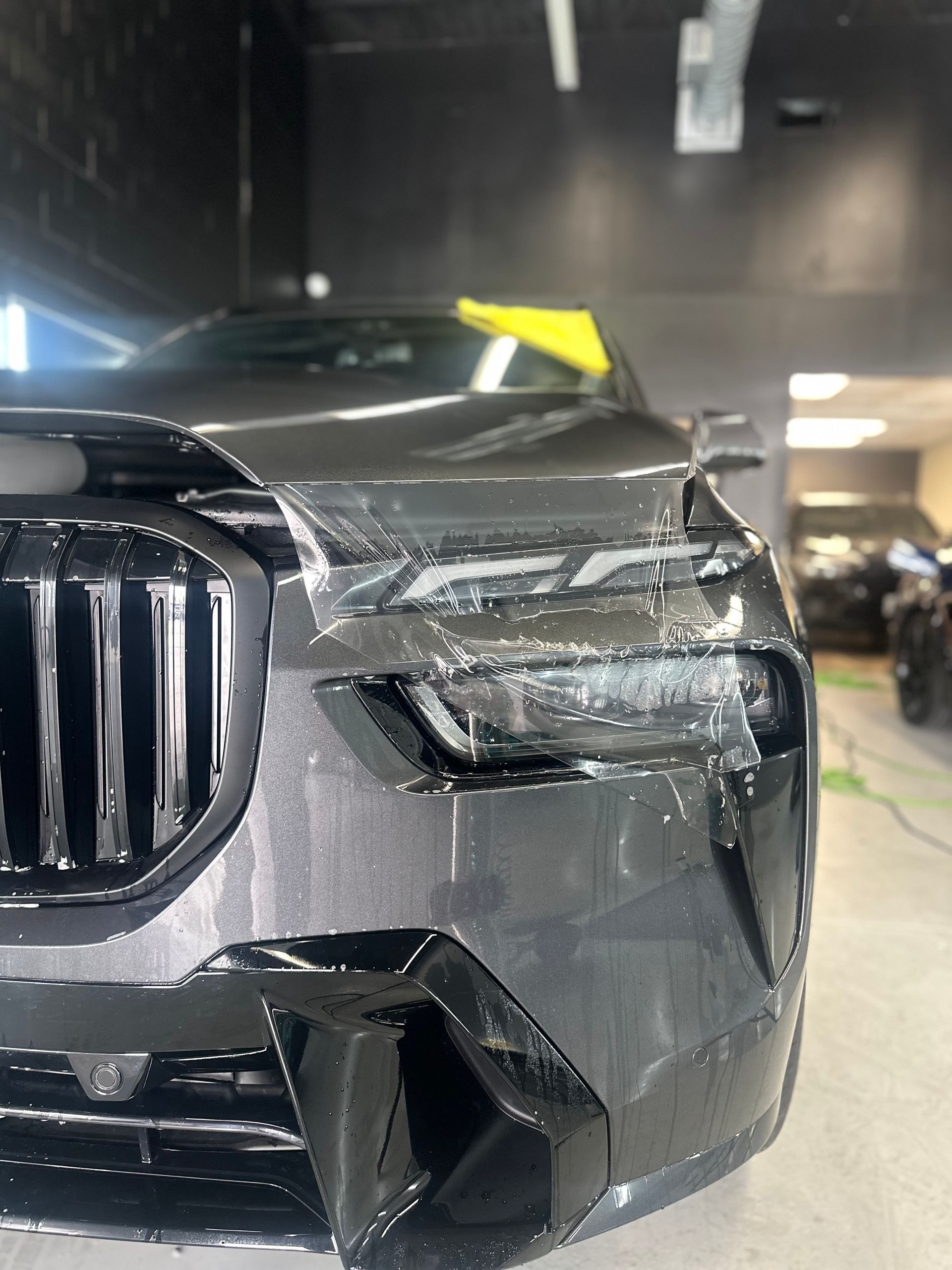 Close-up of a dark grey car being covered with transparent paint protection film at a workshop.