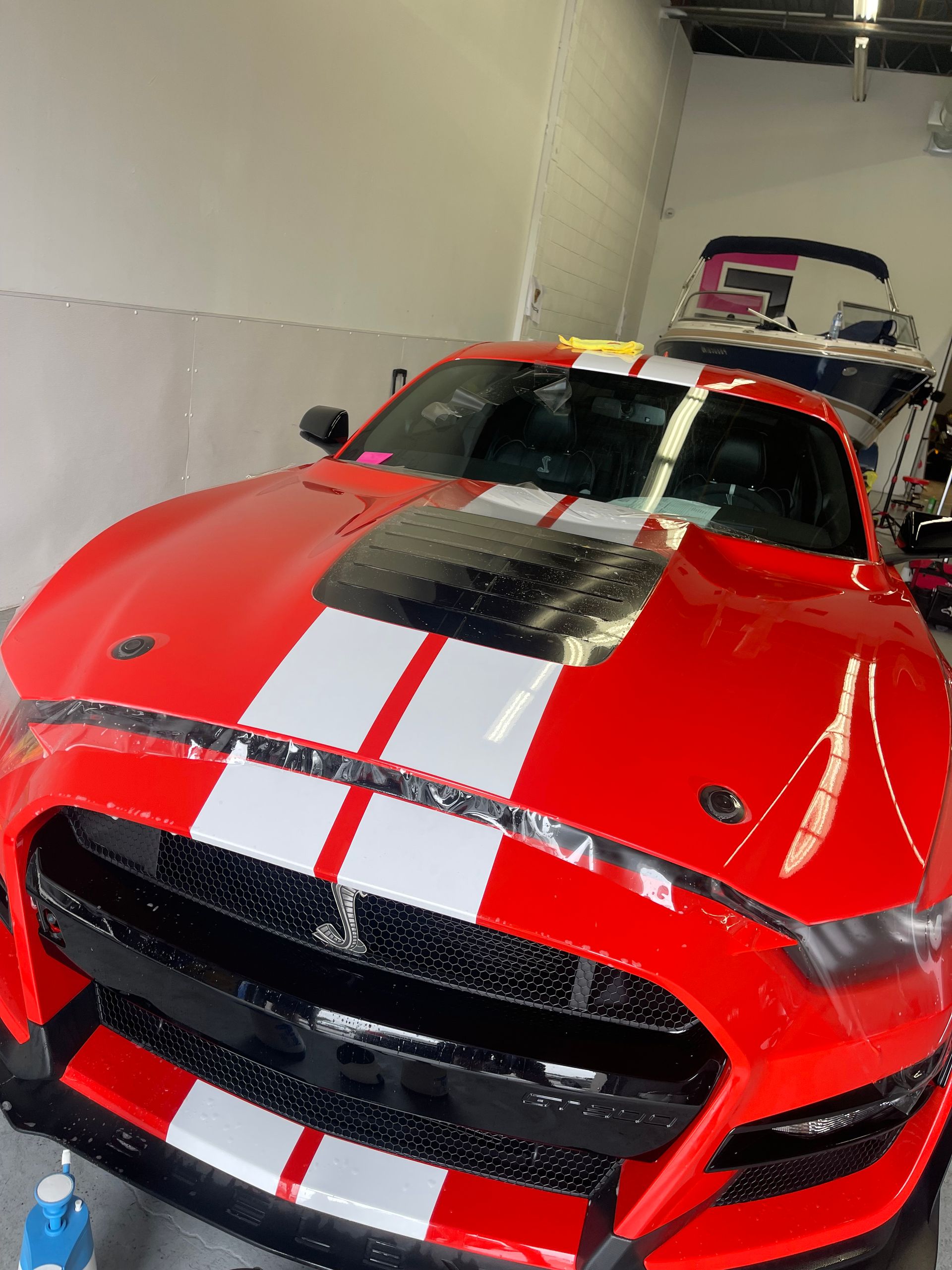 A bright red Ford Mustang with white racing stripes parked in a garage with a boat visible in the background.