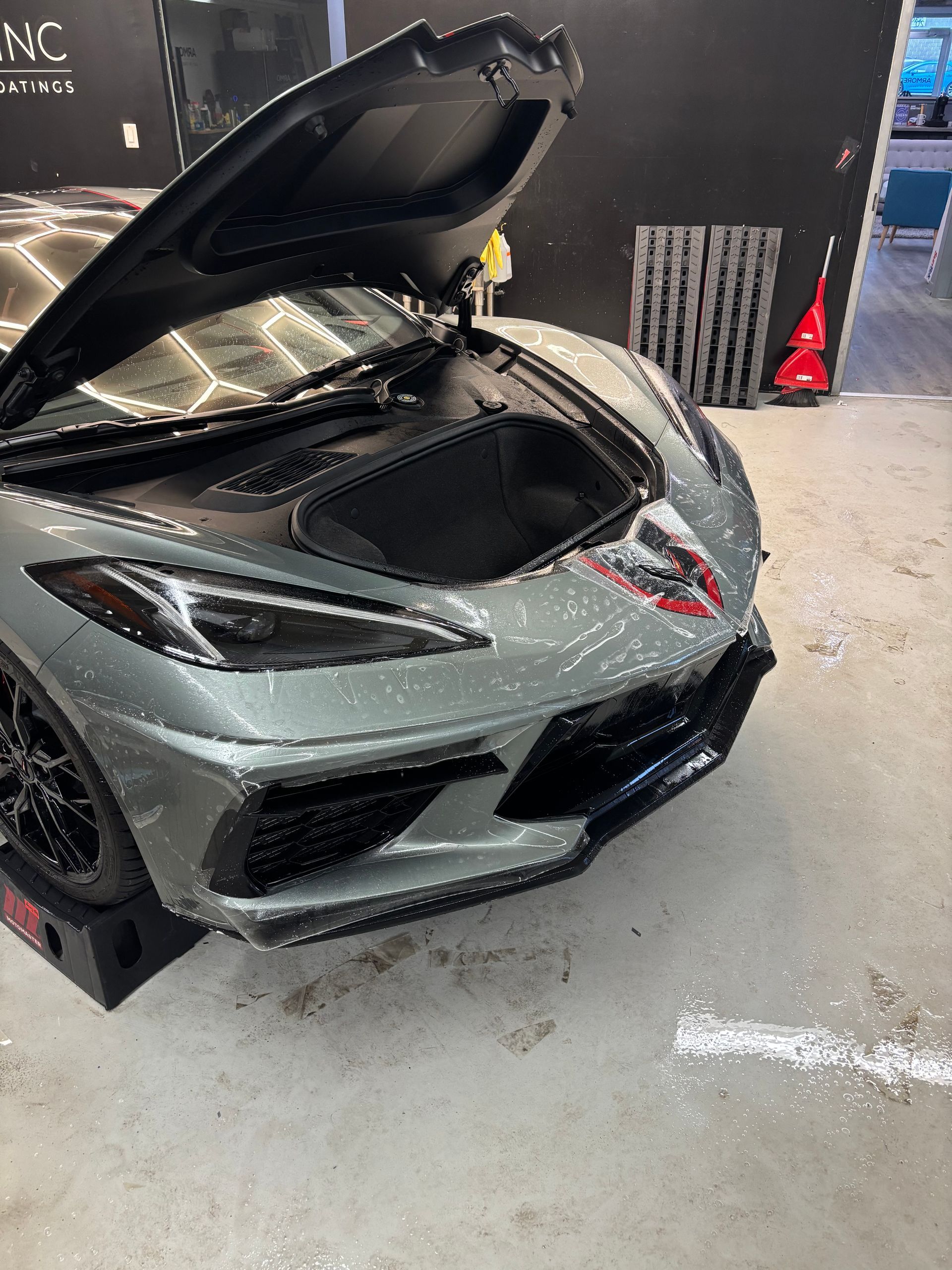 A grey C8 Chevrolet Corvette parked in a garage with its front trunk hood open and parked on black ramps.
