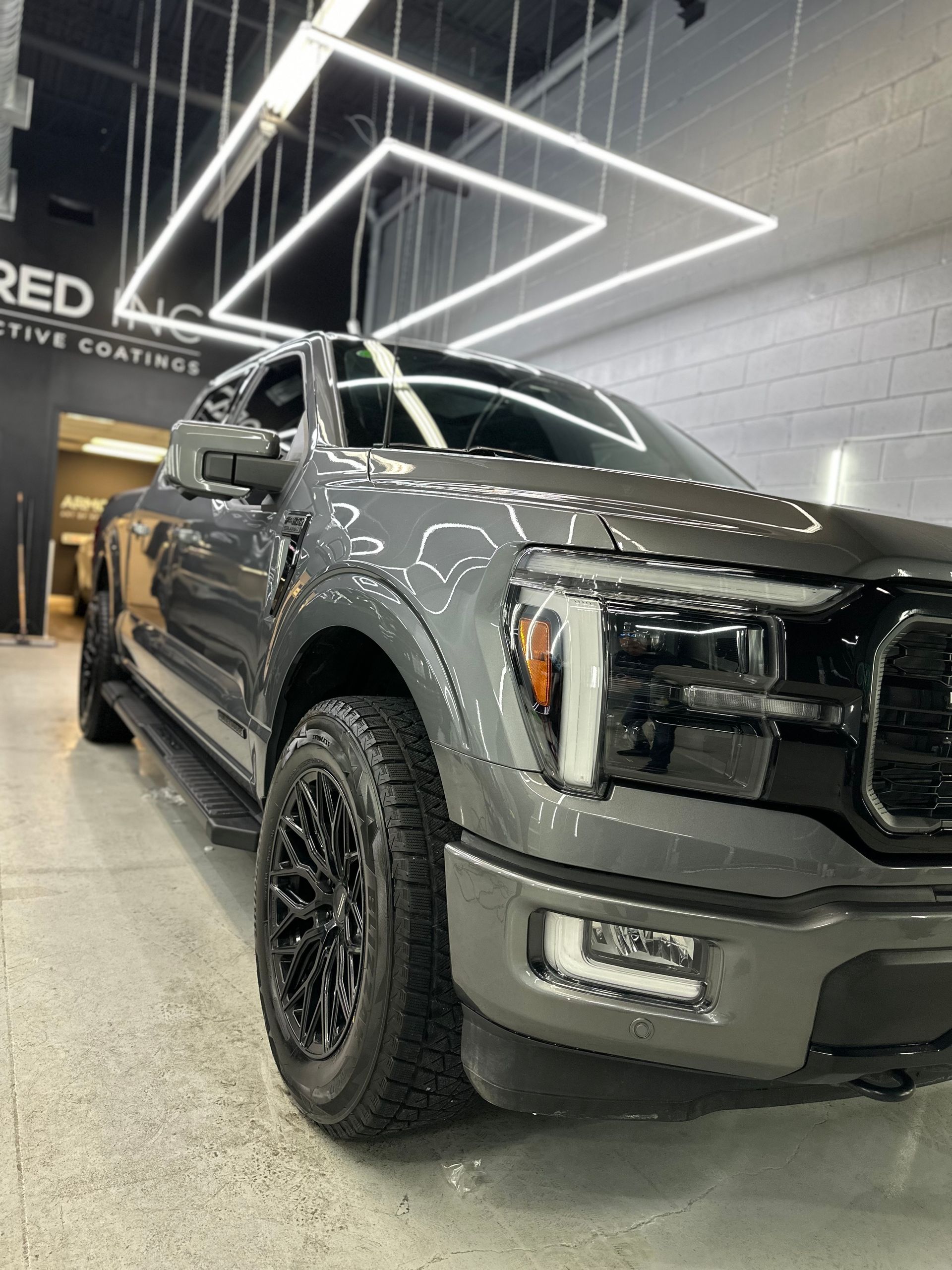 A dark grey Ford F-150 pickup truck parked inside a bright, modern auto shop with overhead geometric lighting.