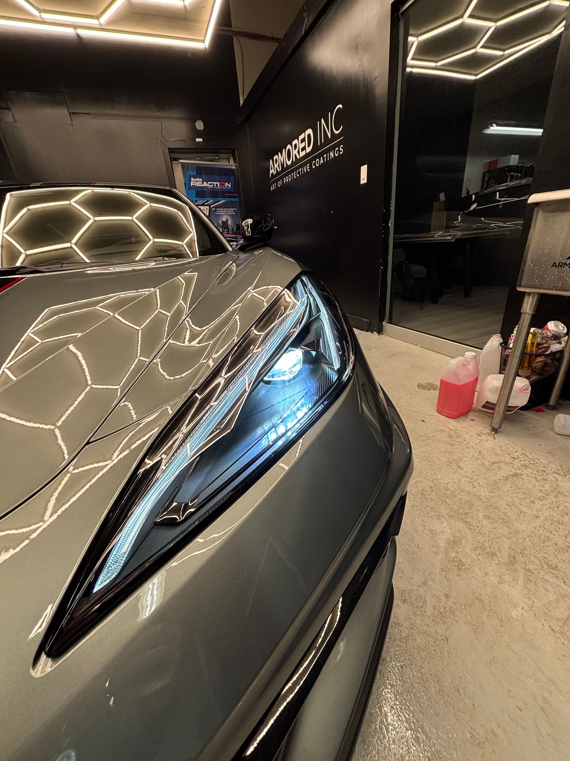A close-up of a grey car's sleek headlight in a dimly lit garage with hexagonal ceiling lights reflecting on the hood.