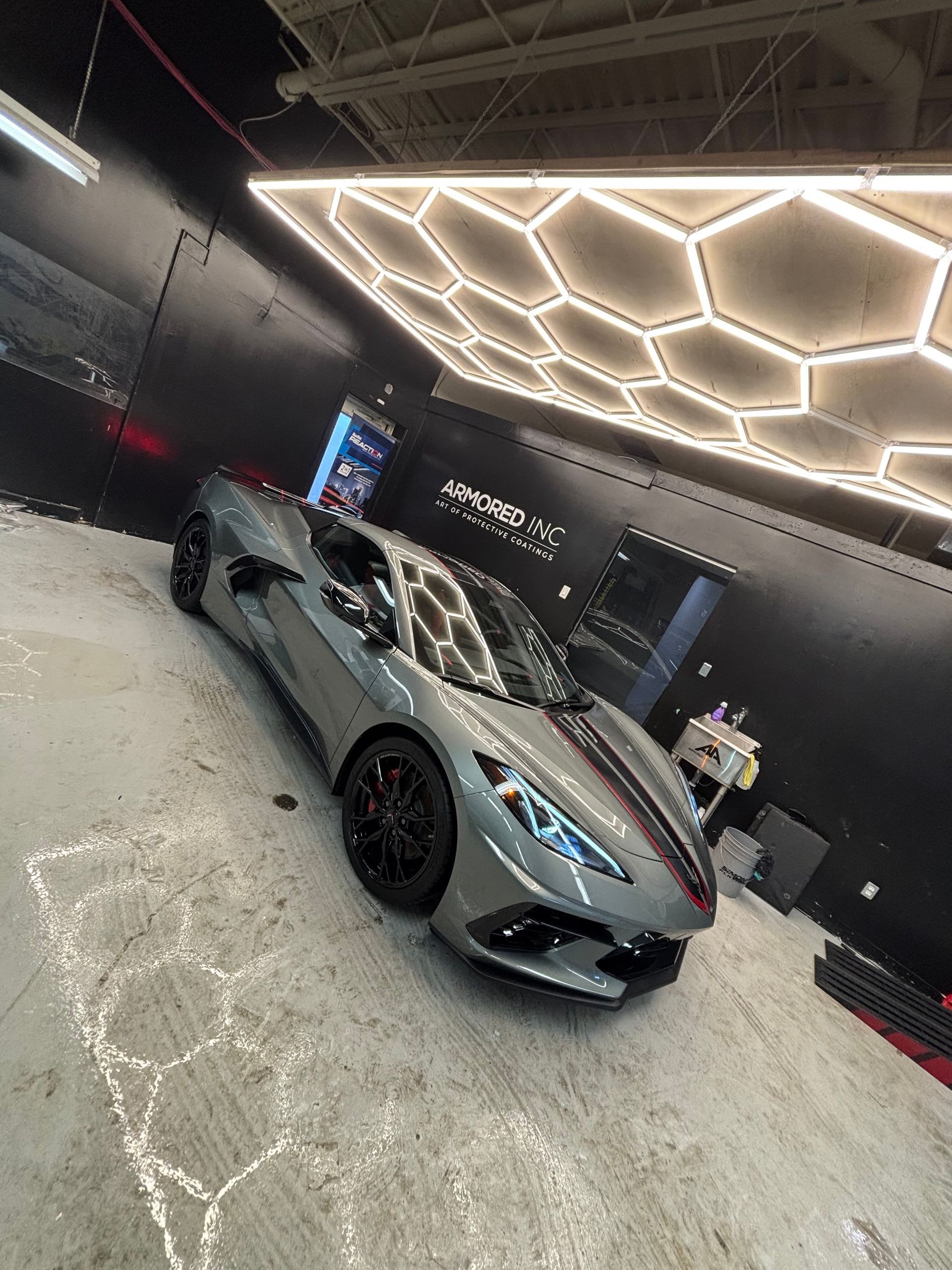 A grey Chevrolet Corvette C8 with a racing stripe parked inside a studio with hexagonal LED ceiling lights.