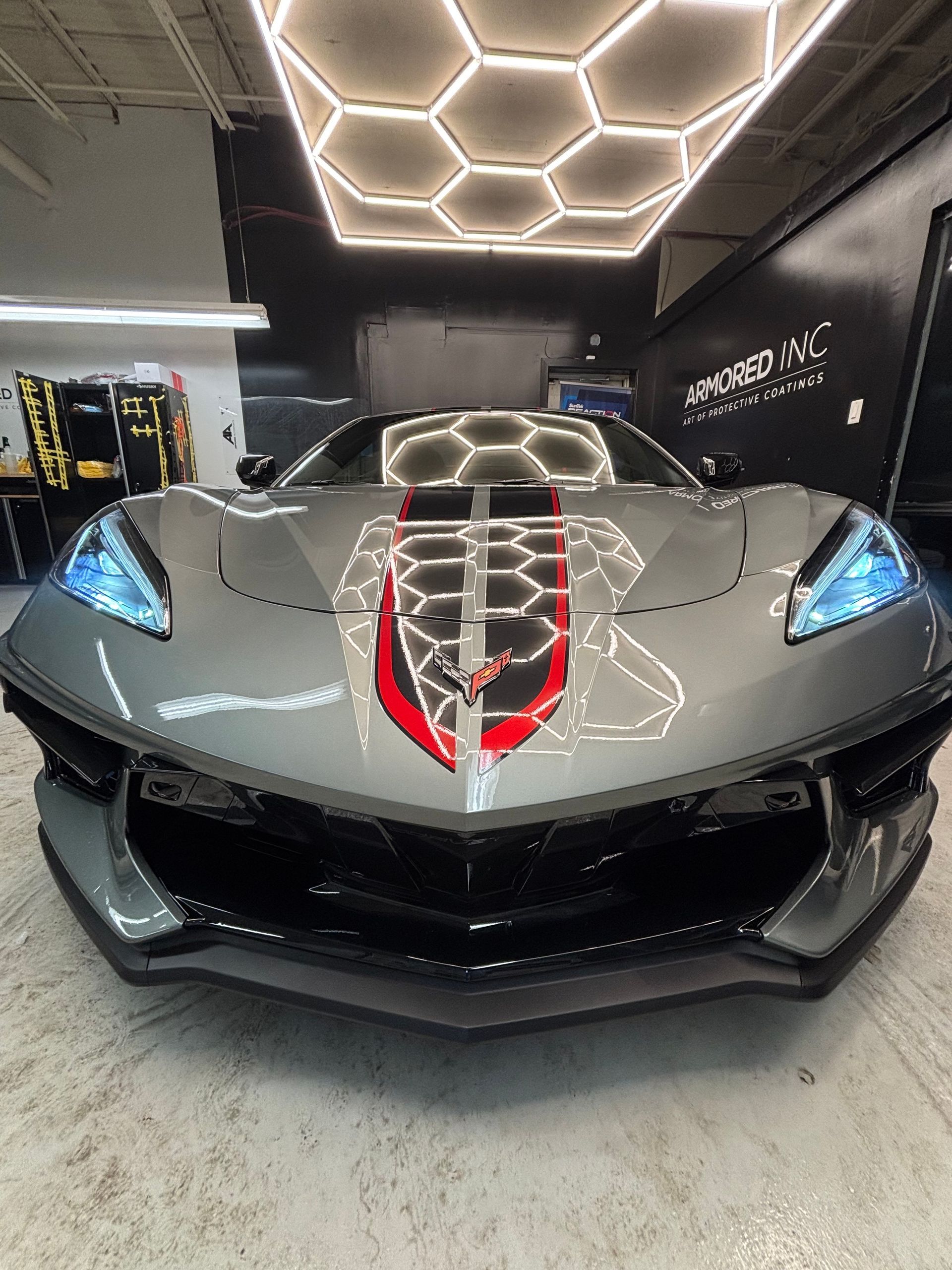 A gray Chevrolet Corvette C8 with a red and black center stripe, parked in a garage with hexagonal overhead lighting.