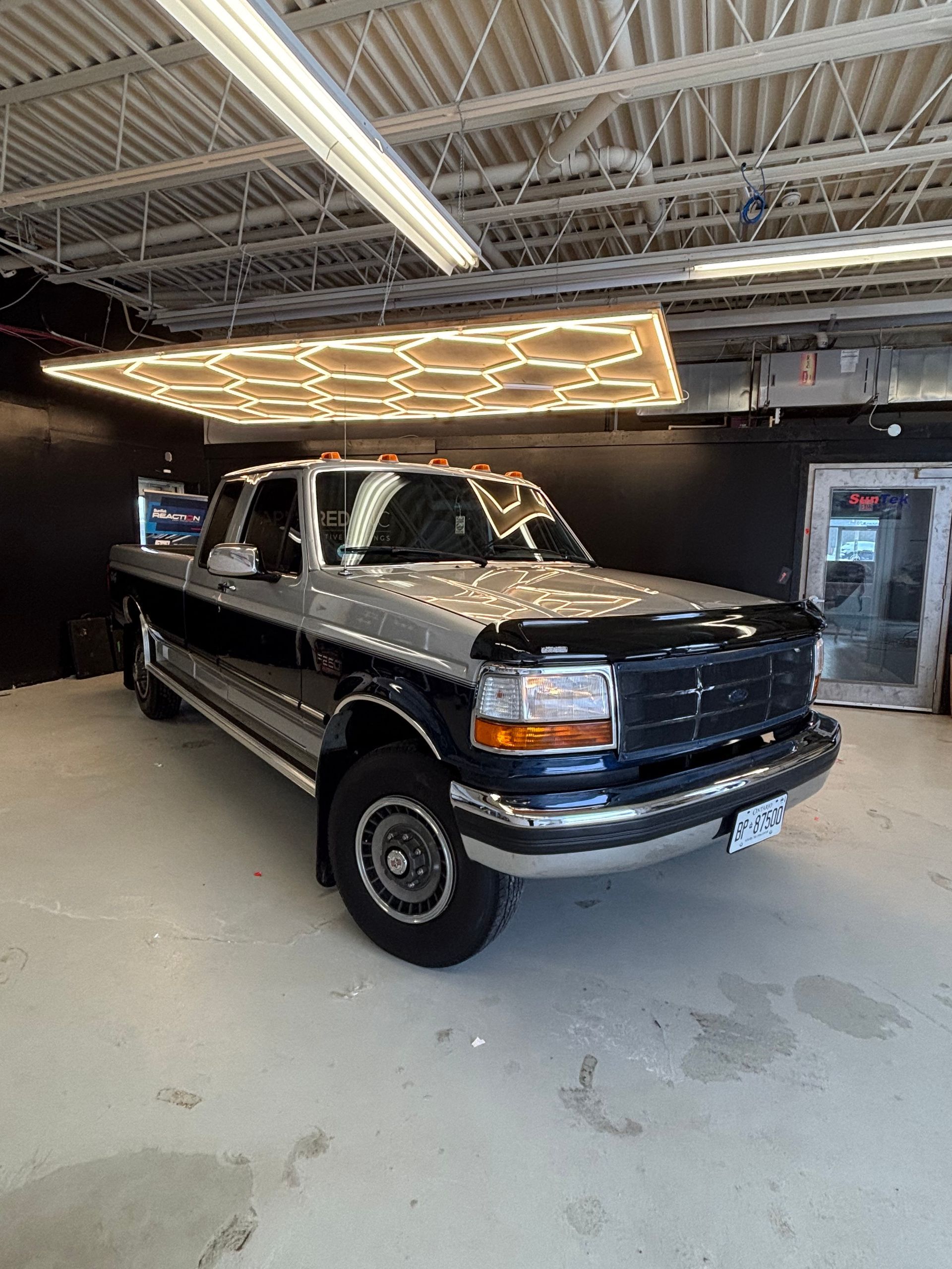 A two-tone silver and black Ford pickup truck parked in a well-lit workshop with hexagonal ceiling lights.