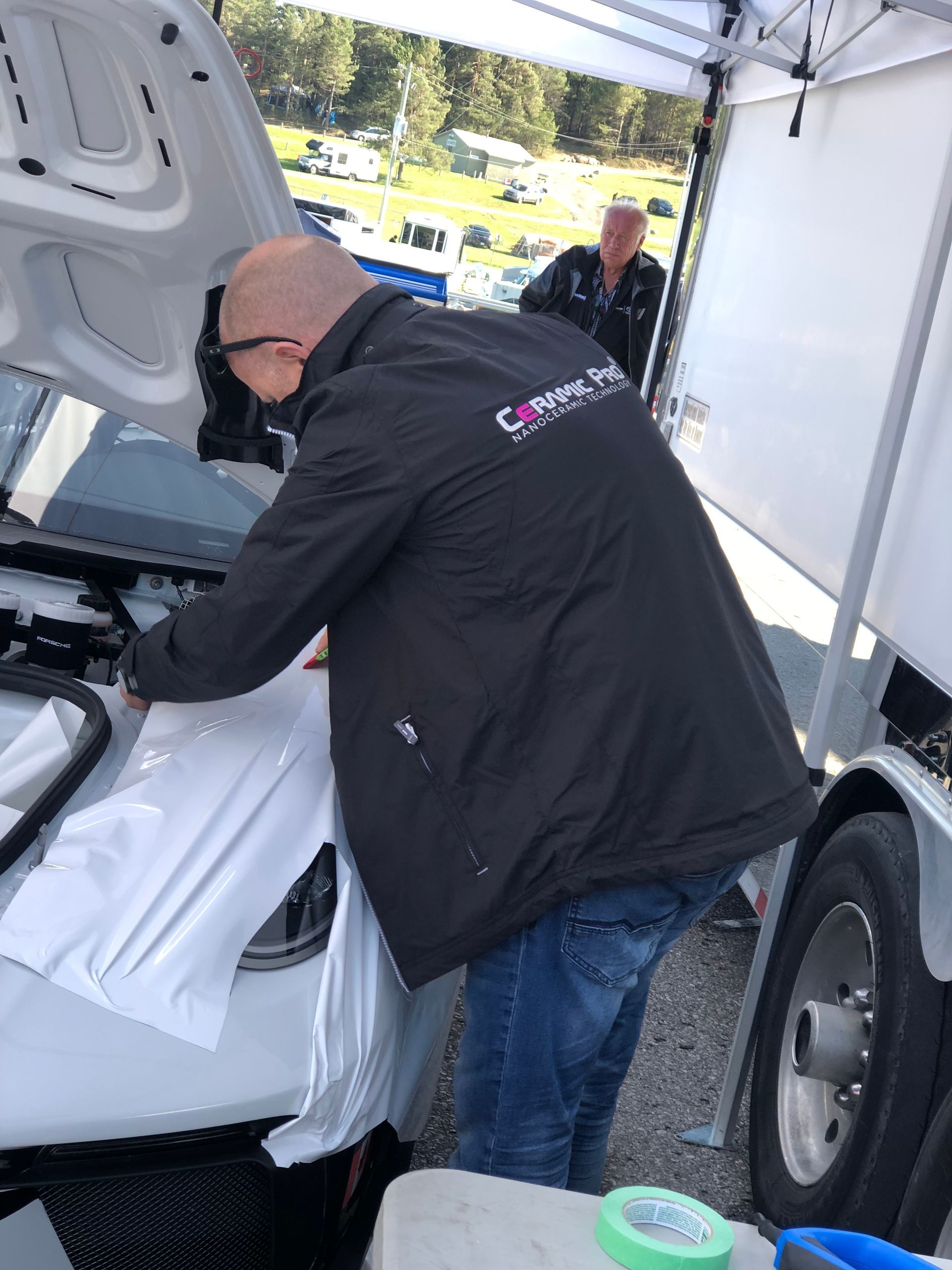 A person applies a white wrap to the front of a vehicle at an outdoor event under a canopy.