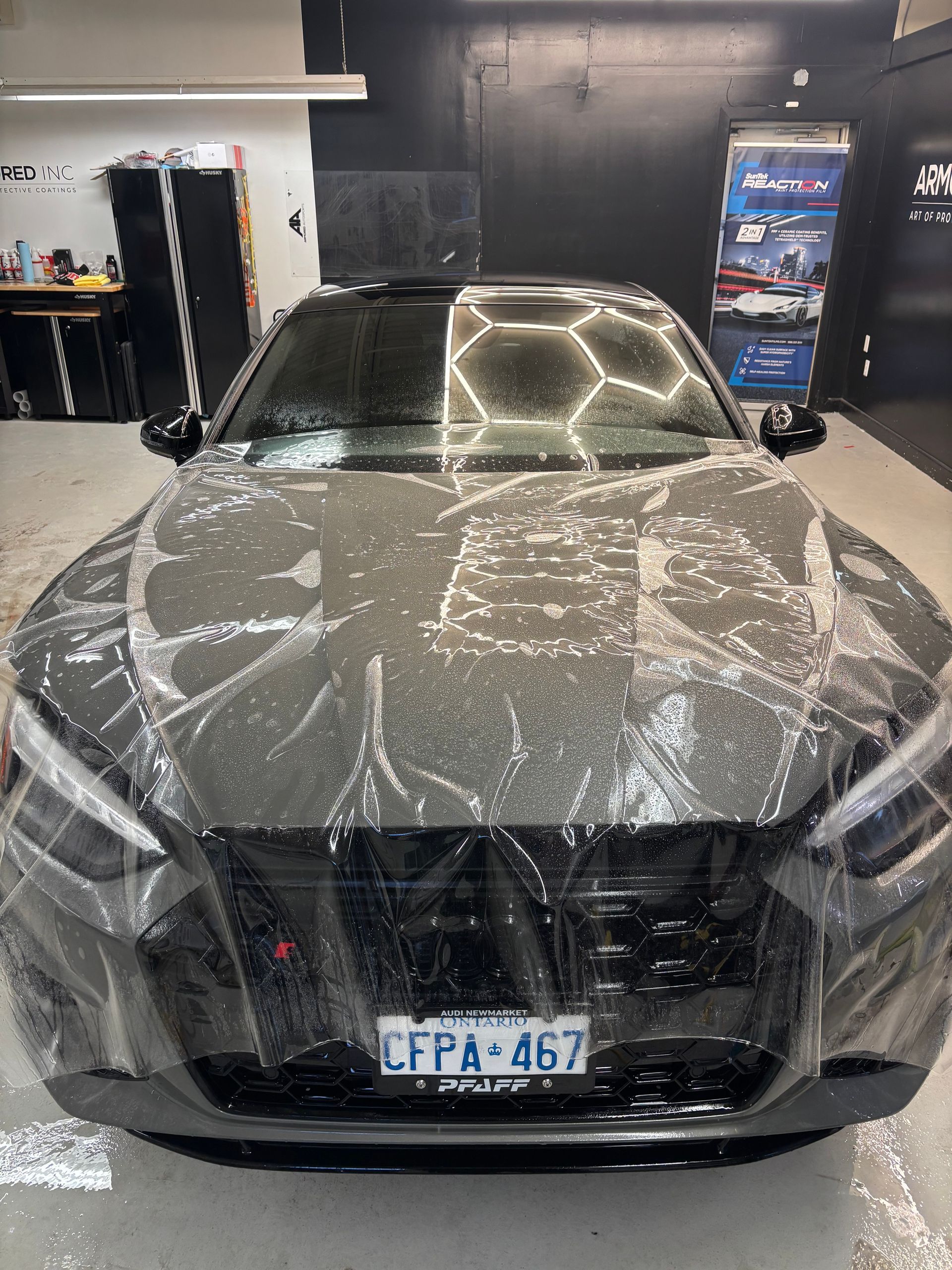 A dark grey car in a garage with clear protective film partially applied across its front hood and bumper.