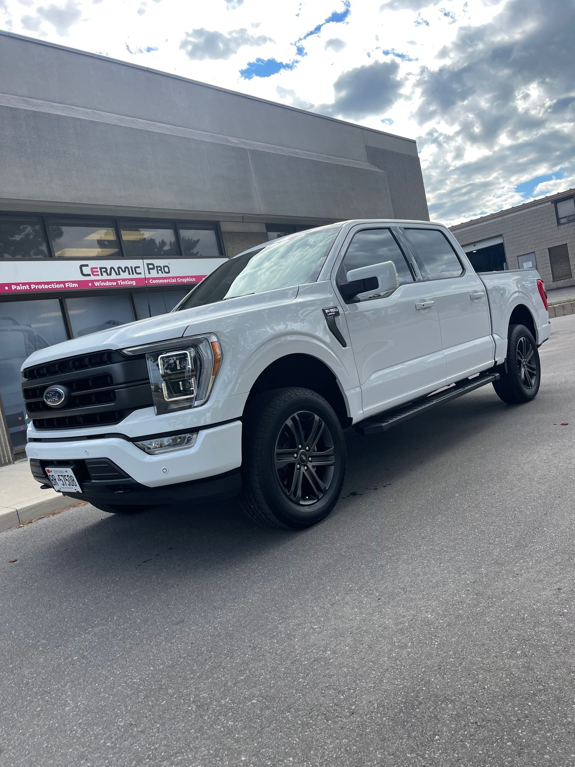 A white Ford F-150 pickup truck with black trim parked in front of a building under a partly cloudy sky.