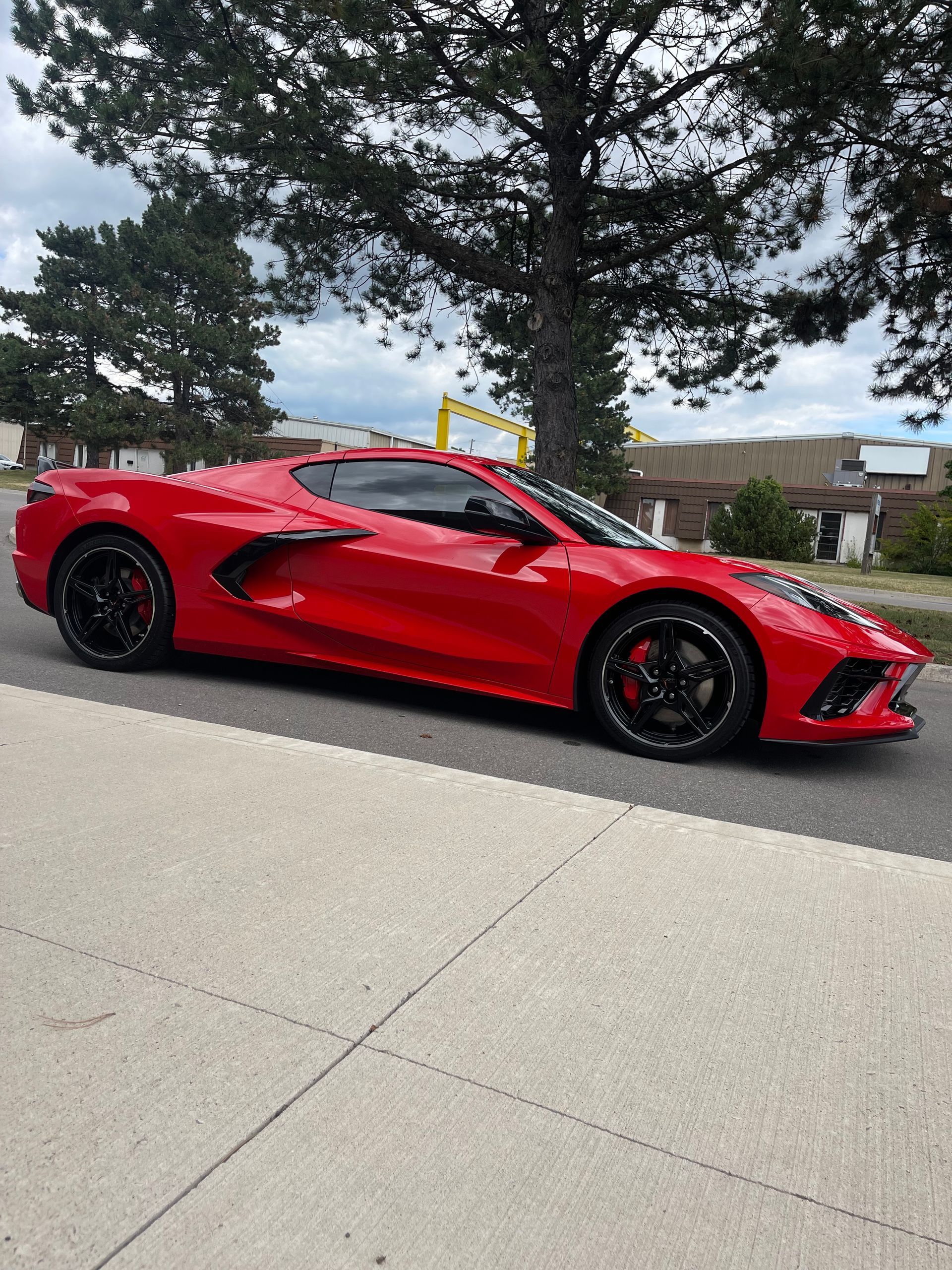 A bright red Chevrolet Corvette sports car parked on an asphalt road in front of a tree and building.