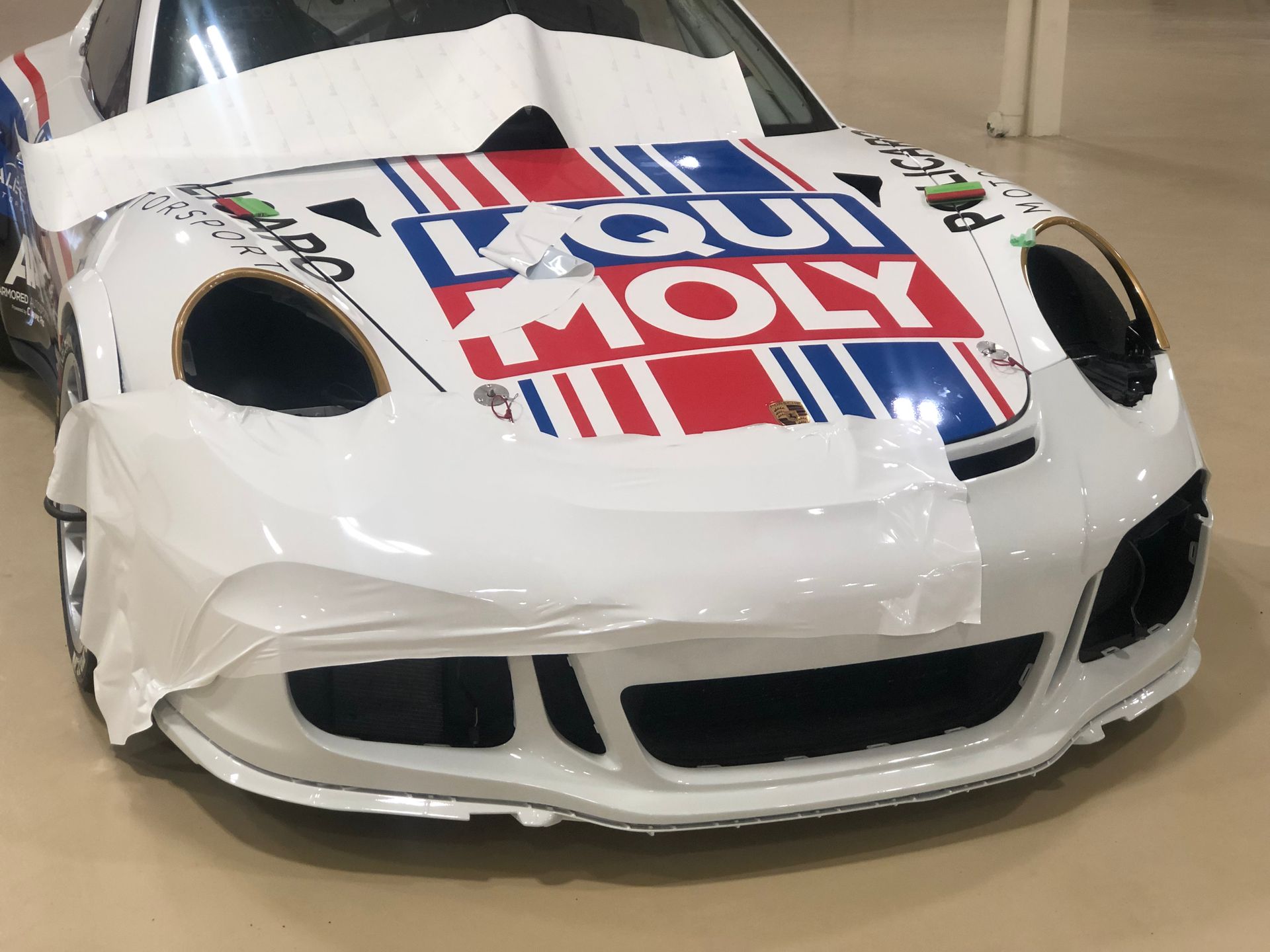 A white Porsche race car with Liqui Moly branding on the hood, partially covered with white protective plastic film.