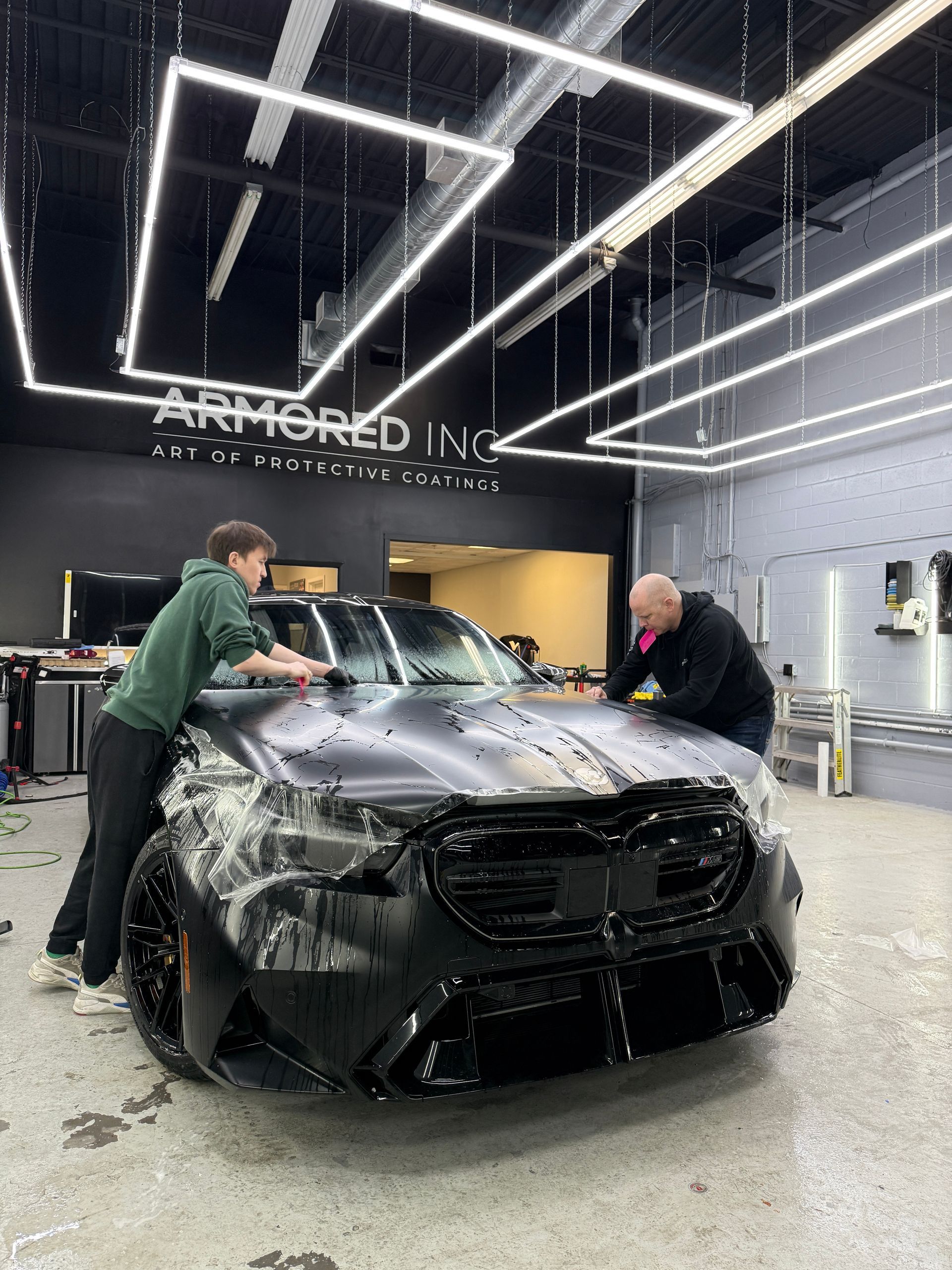 Two technicians apply a protective film to the hood of a black car inside a well-lit auto detailing shop.