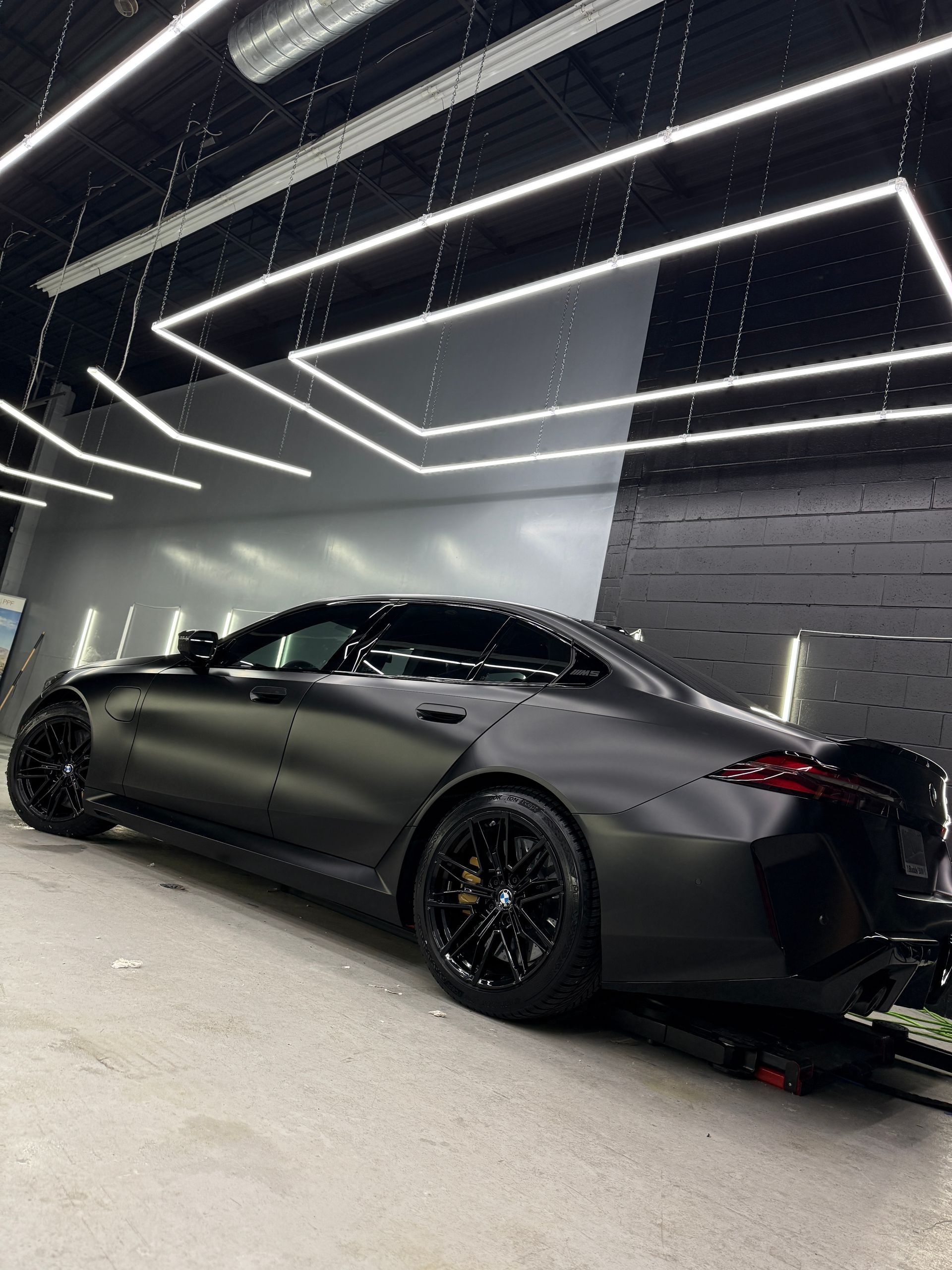 A matte black sedan parked in a studio with modern overhead lighting and a textured black and grey wall.