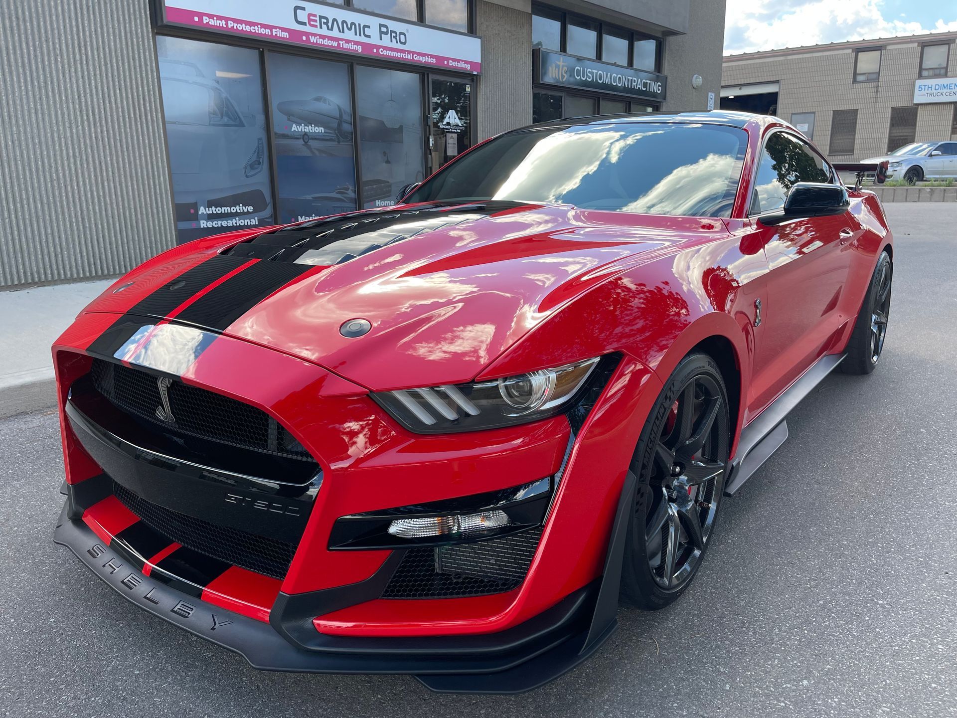 A red Ford Mustang Shelby GT500 with black racing stripes parked in front of an automotive shop.