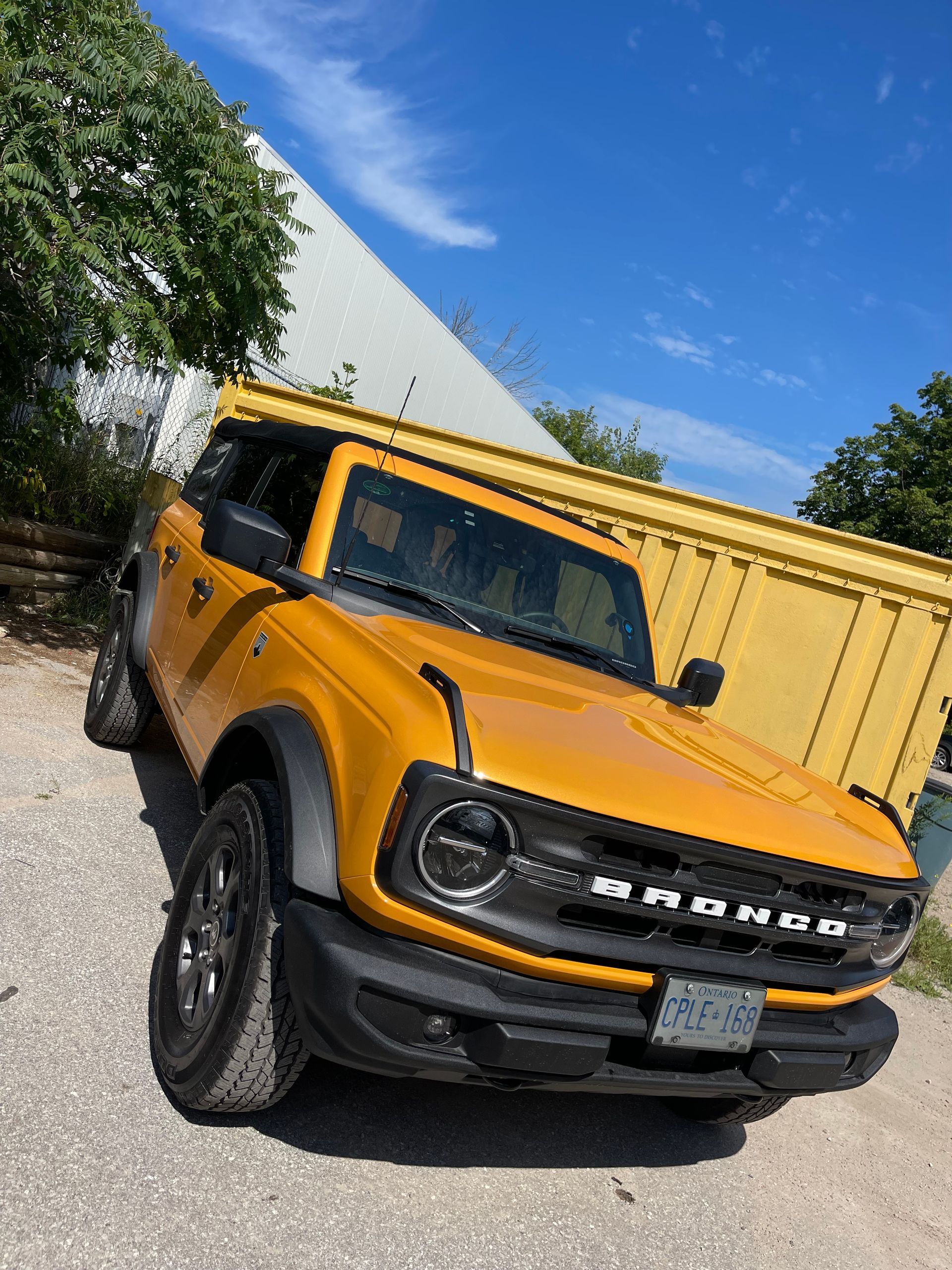 A yellow Ford Bronco parked on a gravel lot next to a large yellow industrial container under a clear blue sky.