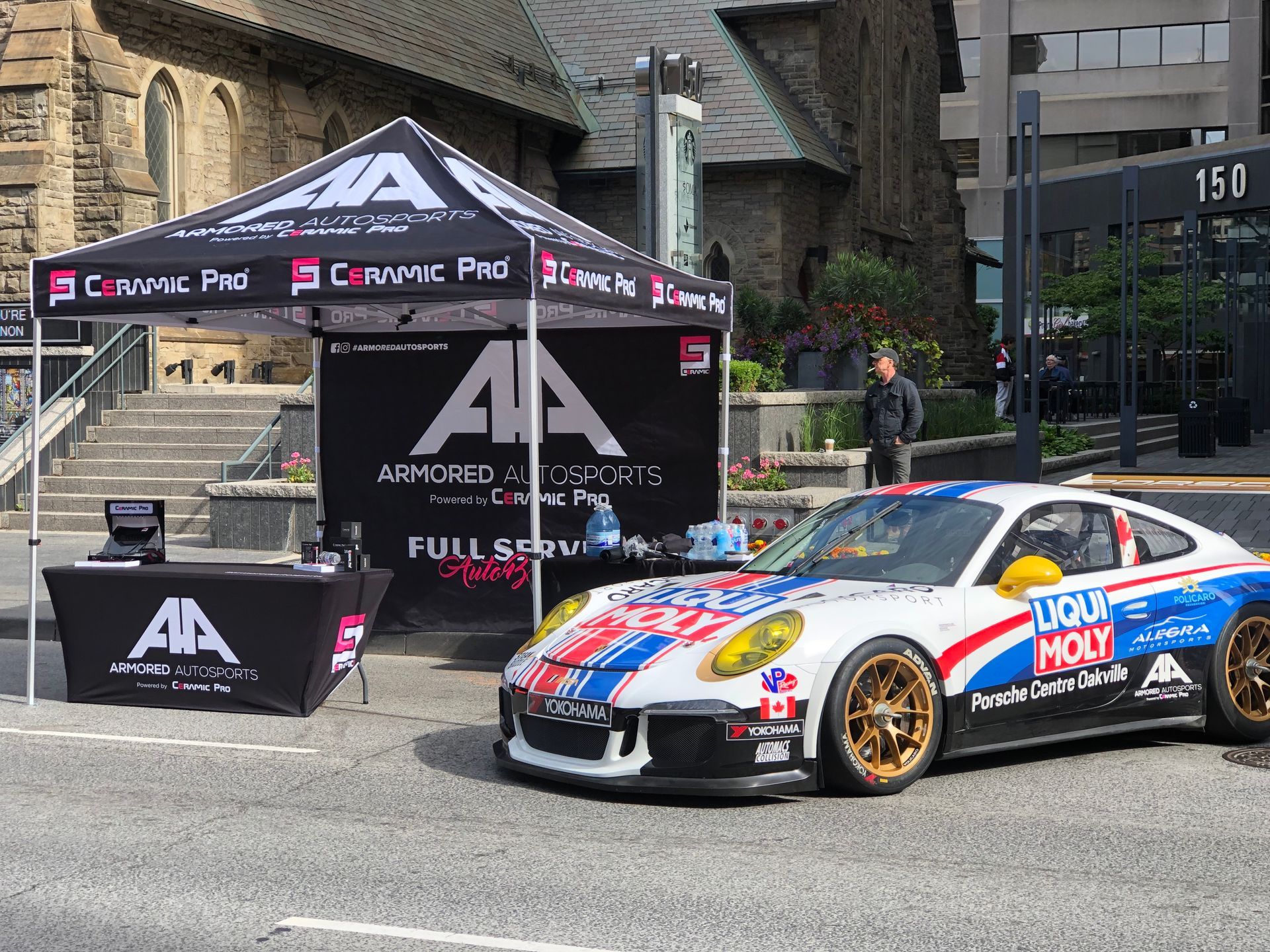 A branded tent with an Armored AutoSports logo next to a white, red, and blue racing Porsche parked on an asphalt street.