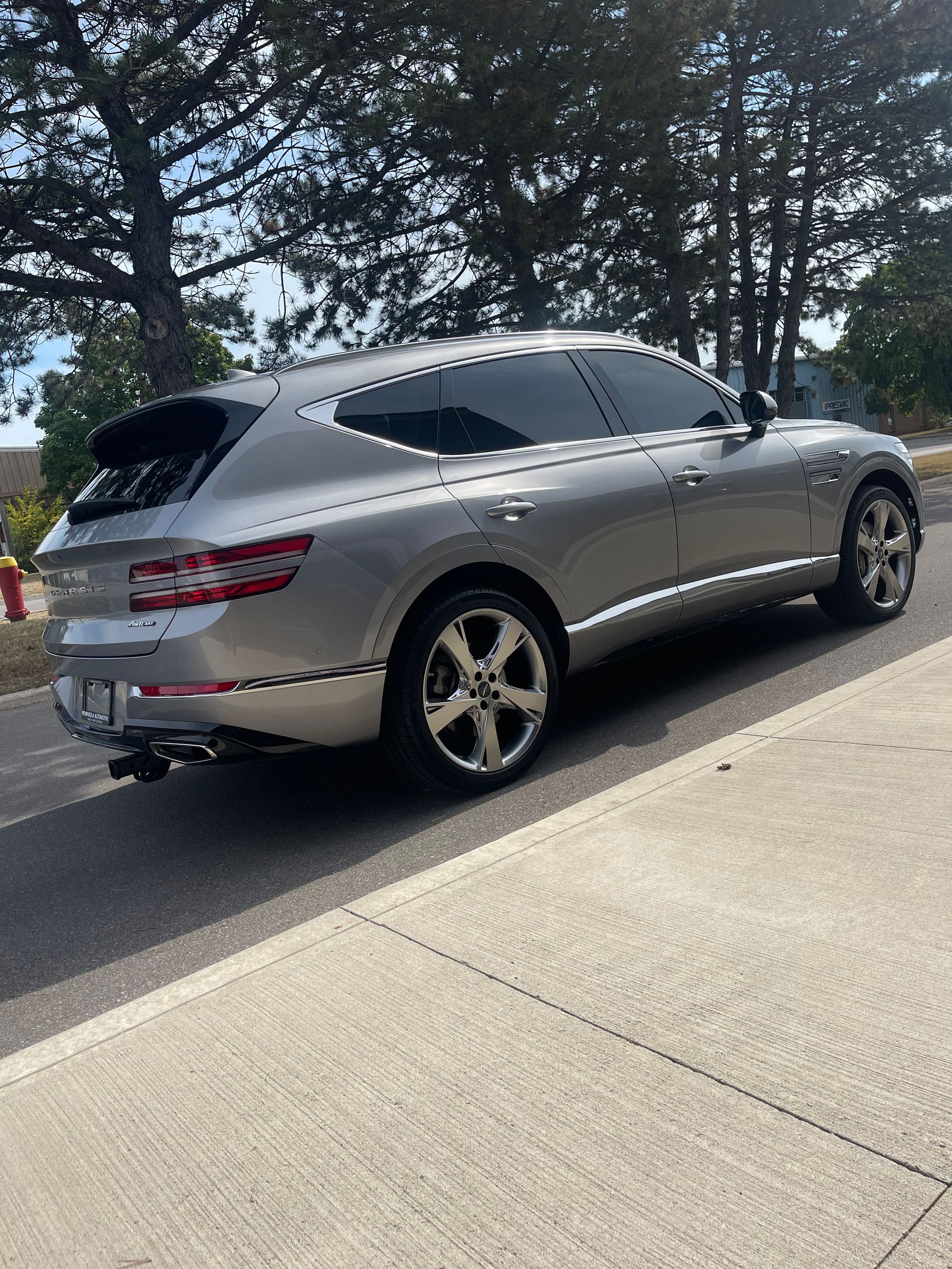 A silver Genesis GV80 SUV parked on an asphalt street next to a concrete sidewalk, with trees in the background.