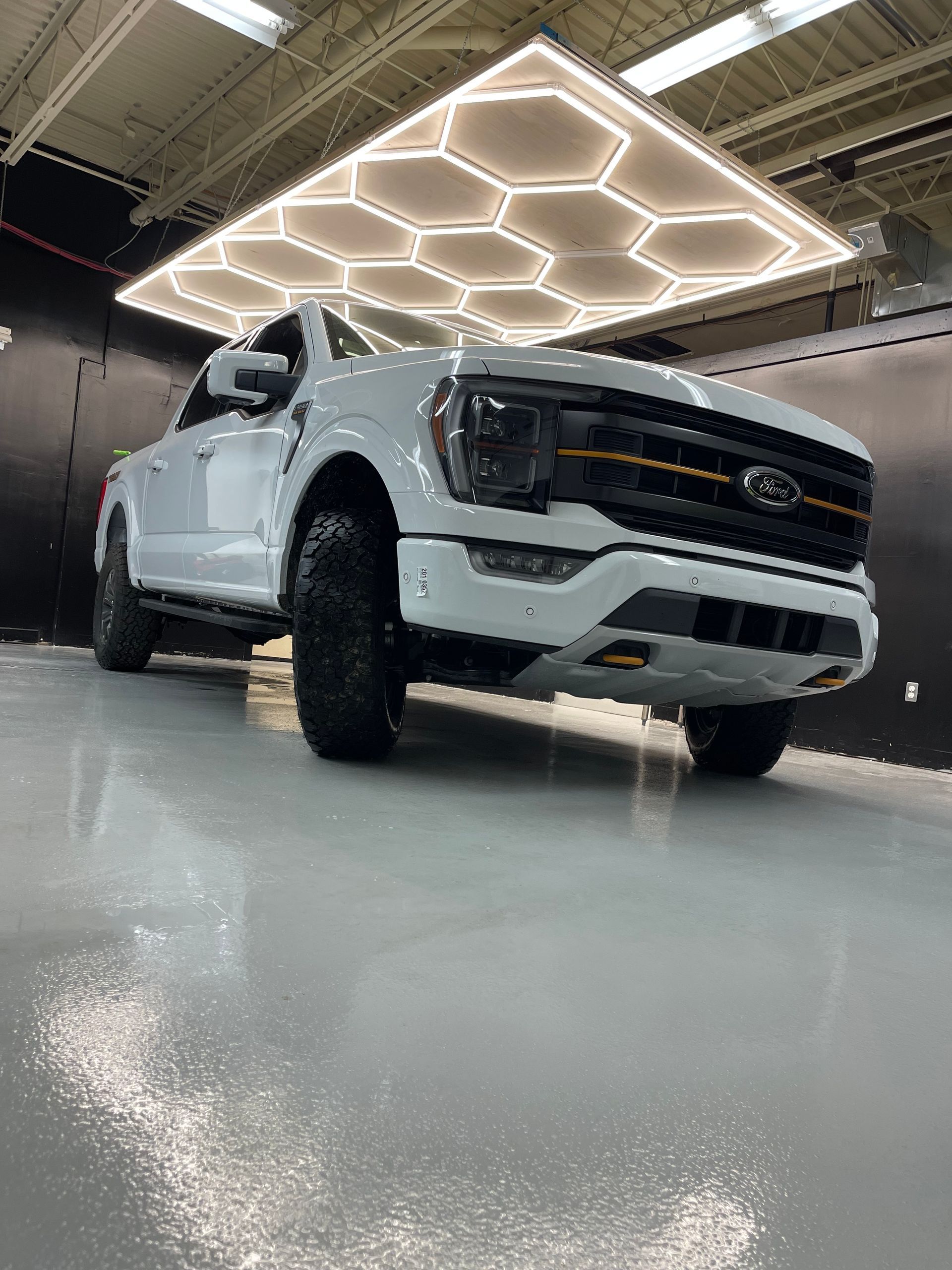A white Ford F-150 truck with orange accents parked on a glossy concrete floor under a honeycomb-patterned LED light.