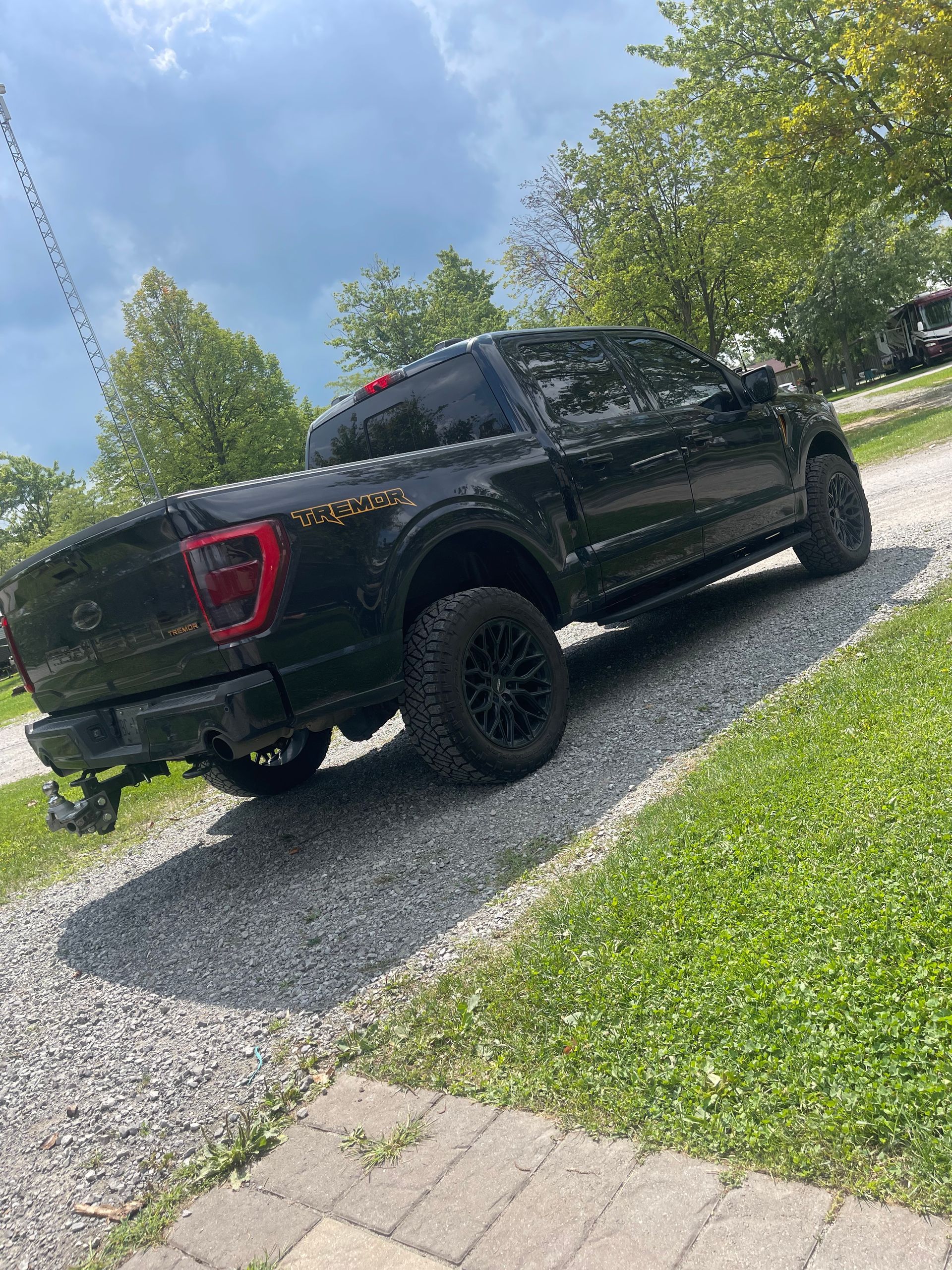 A black Ford F-150 pickup truck parked on a gravel driveway next to a grass lawn under a partly cloudy sky.