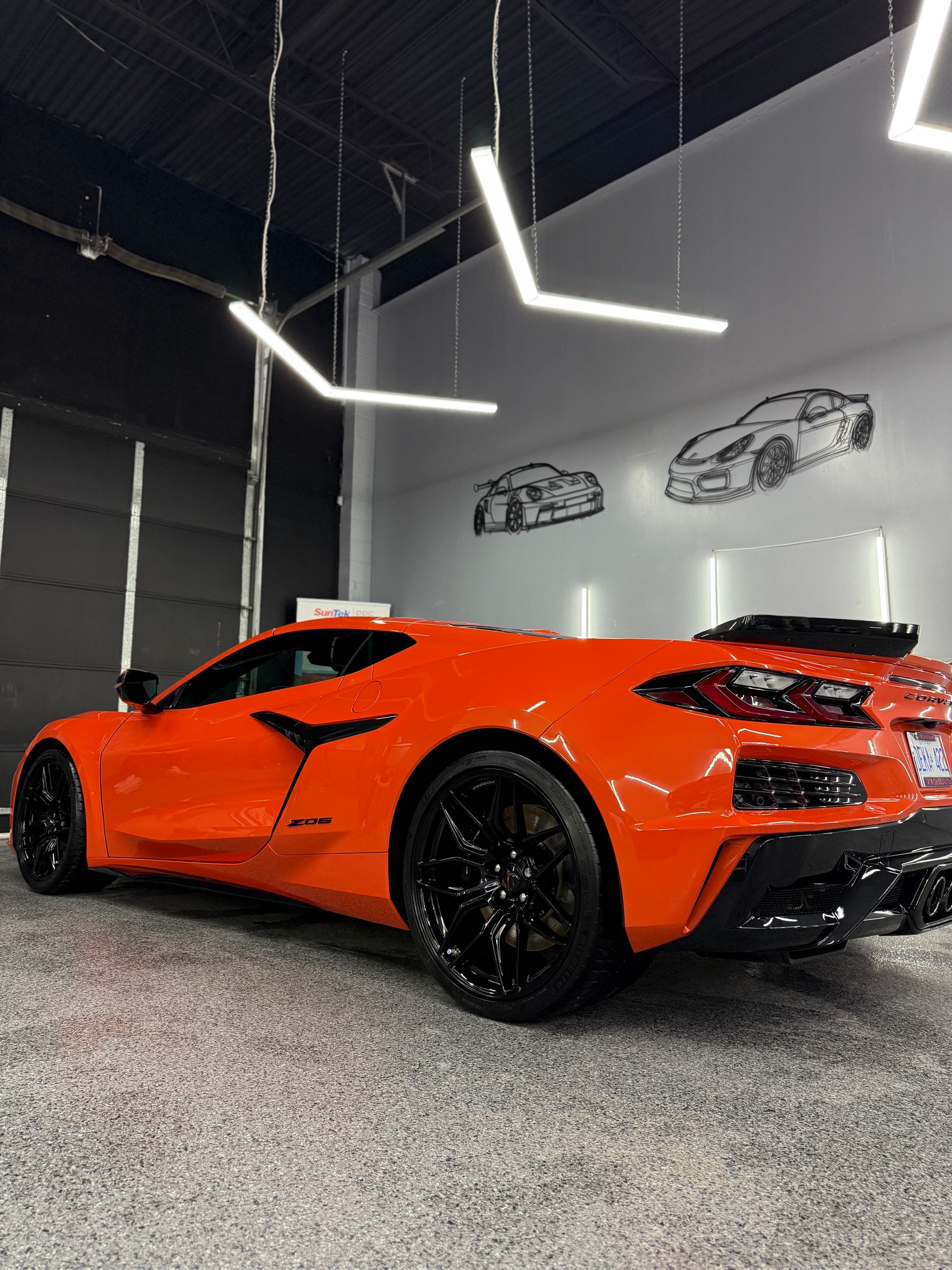 An orange Chevrolet Corvette parked in a modern garage with black wheels and wall-mounted line art of cars.