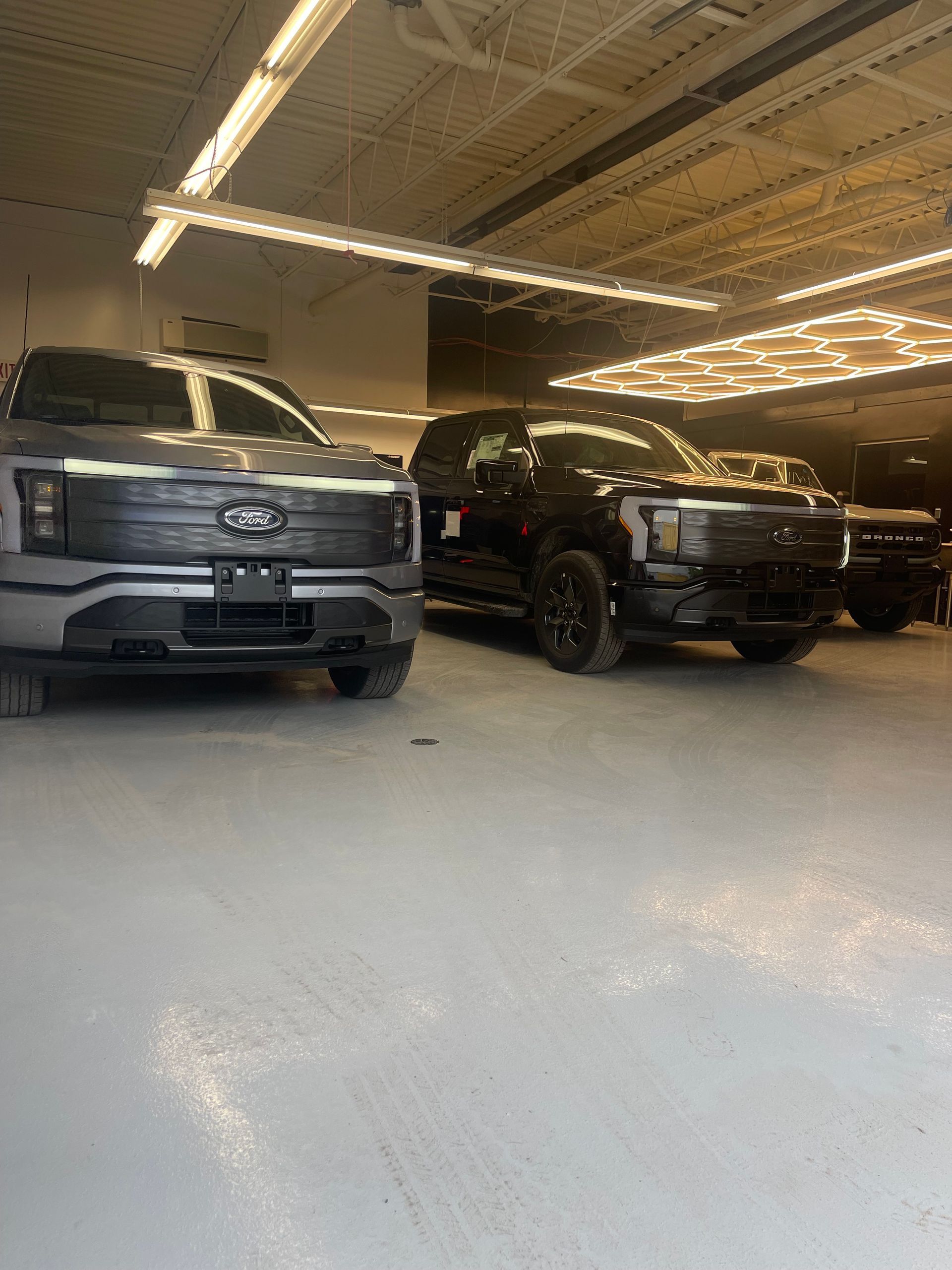 Two Ford F-150 Lightning trucks, one silver and one black, parked inside a well-lit showroom with industrial flooring.