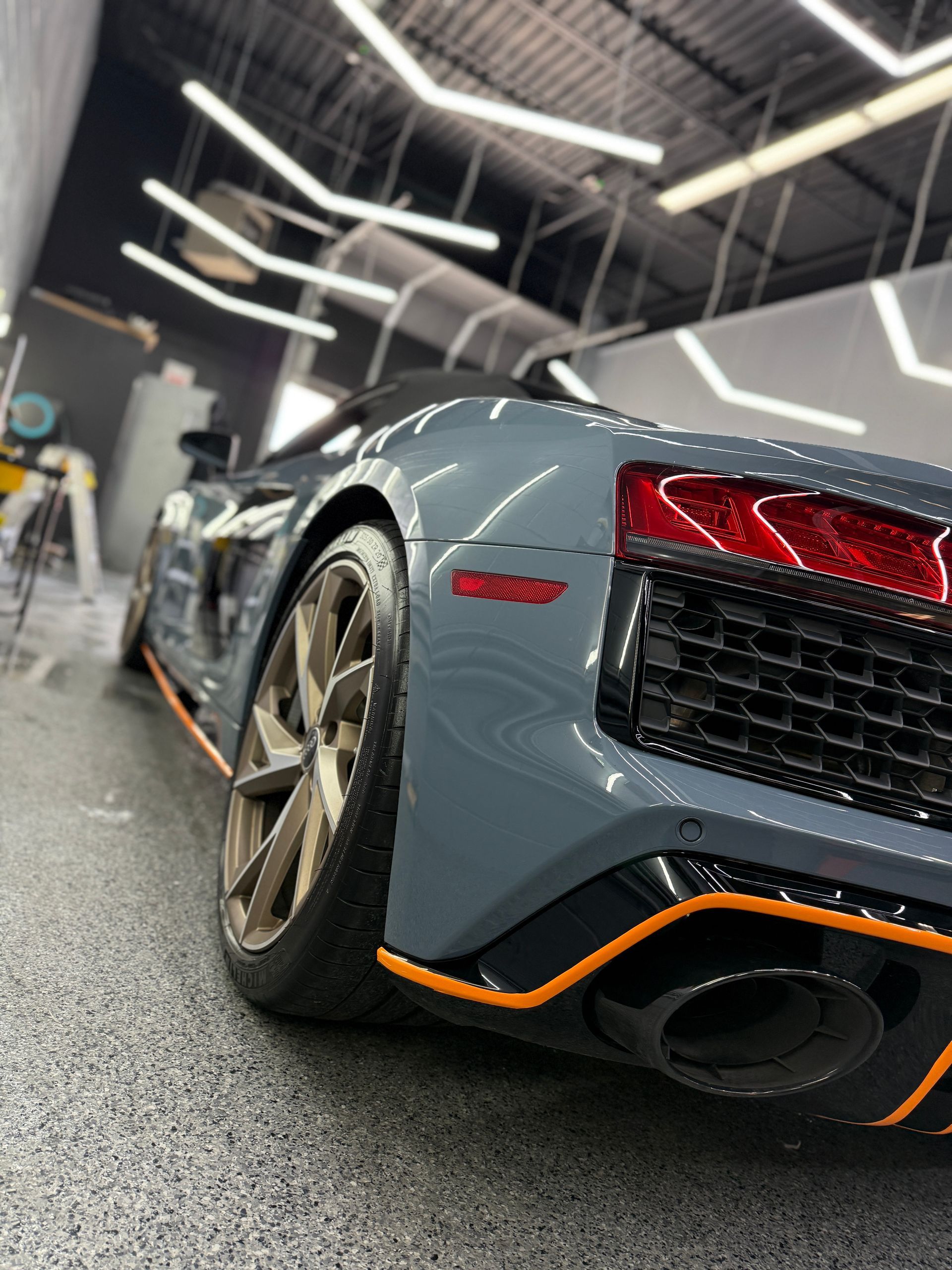 A gray Audi R8 with orange accents parked in a garage with bright, angular overhead lighting.
