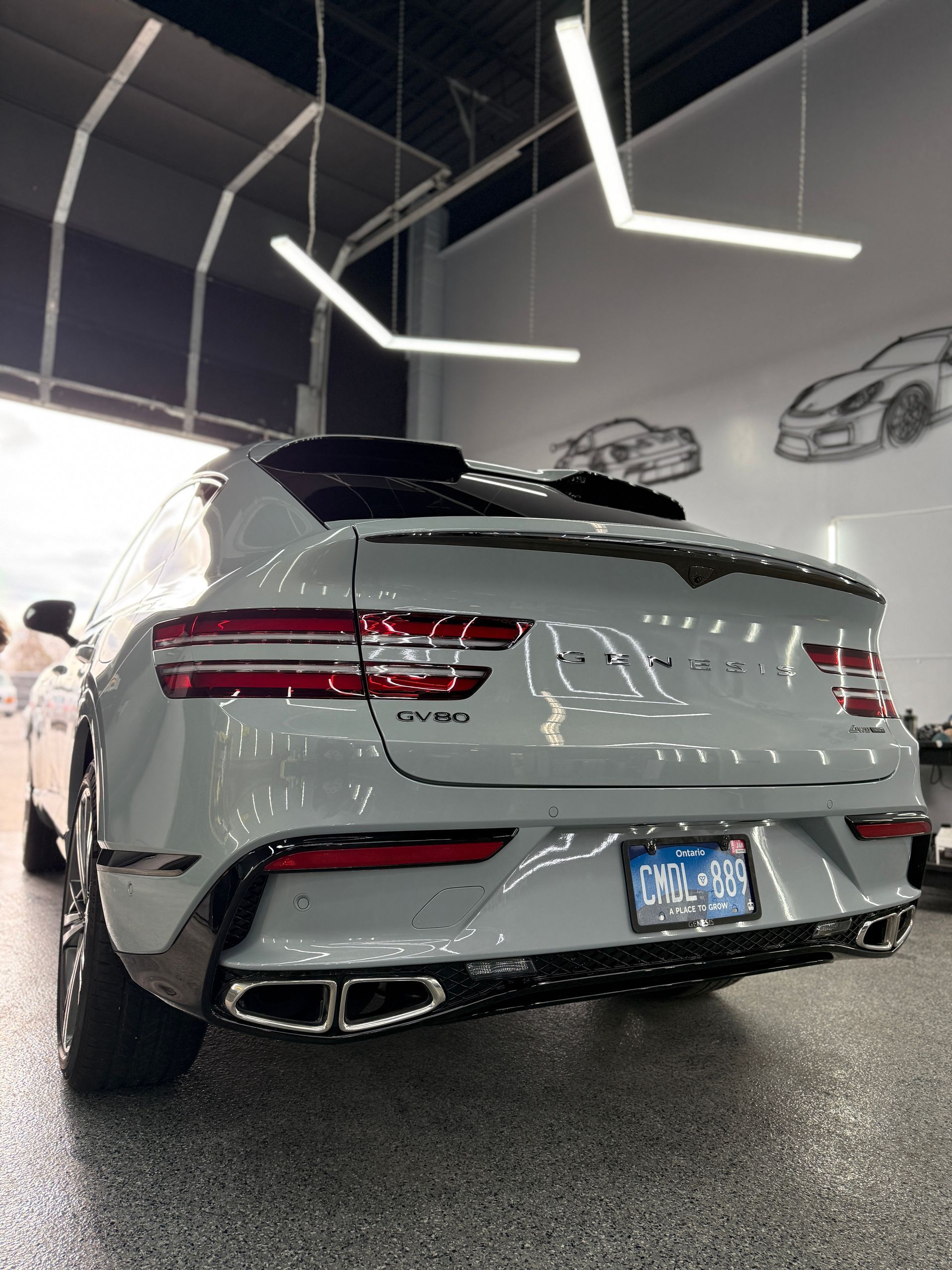 A light gray Genesis GV70 SUV parked in a brightly lit, modern garage with decorative car drawings on the white walls.