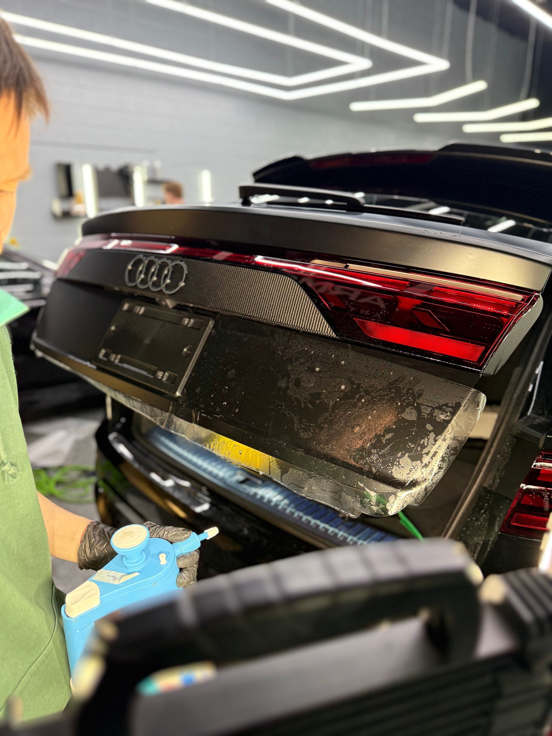A person in a garage sprays liquid onto the open trunk of a black Audi, likely applying protective film or cleaning it.